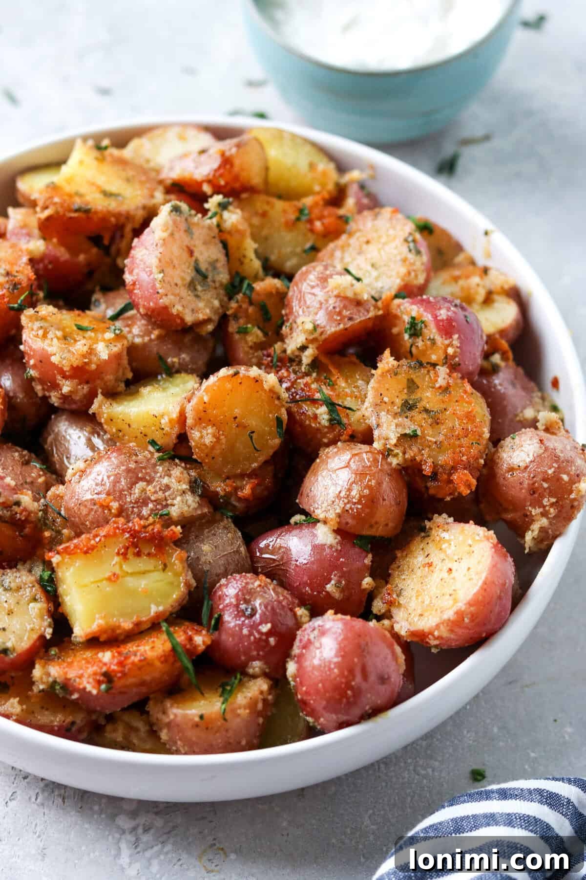 A close-up shot of crispy parmesan potatoes in a white bowl, with a blurry background of sour cream and fresh herbs, suggesting a serving suggestion.