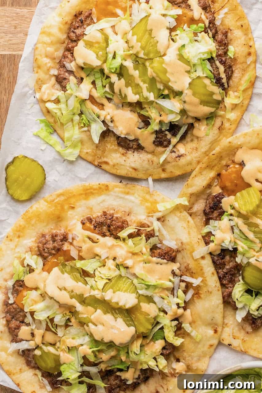 Close-up of smash burger tacos on parchment paper, garnished with shredded lettuce, sliced pickles, melted cheese, and big mac sauce.