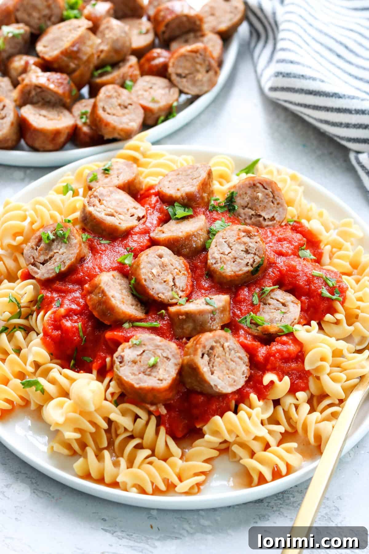 A plate of pasta with marinara sauce and sliced Italian sausage, with a gold fork, and air-fried sausages in the background.