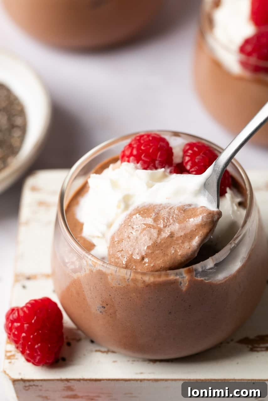 A spoon scooping protein chia seed pudding topped with whipped cream and raspberries from a glass jar.