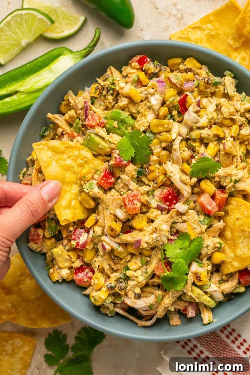 A vibrant bowl of Mexican chicken salad, with a hand dipping a tortilla chip into the creamy, colorful mixture