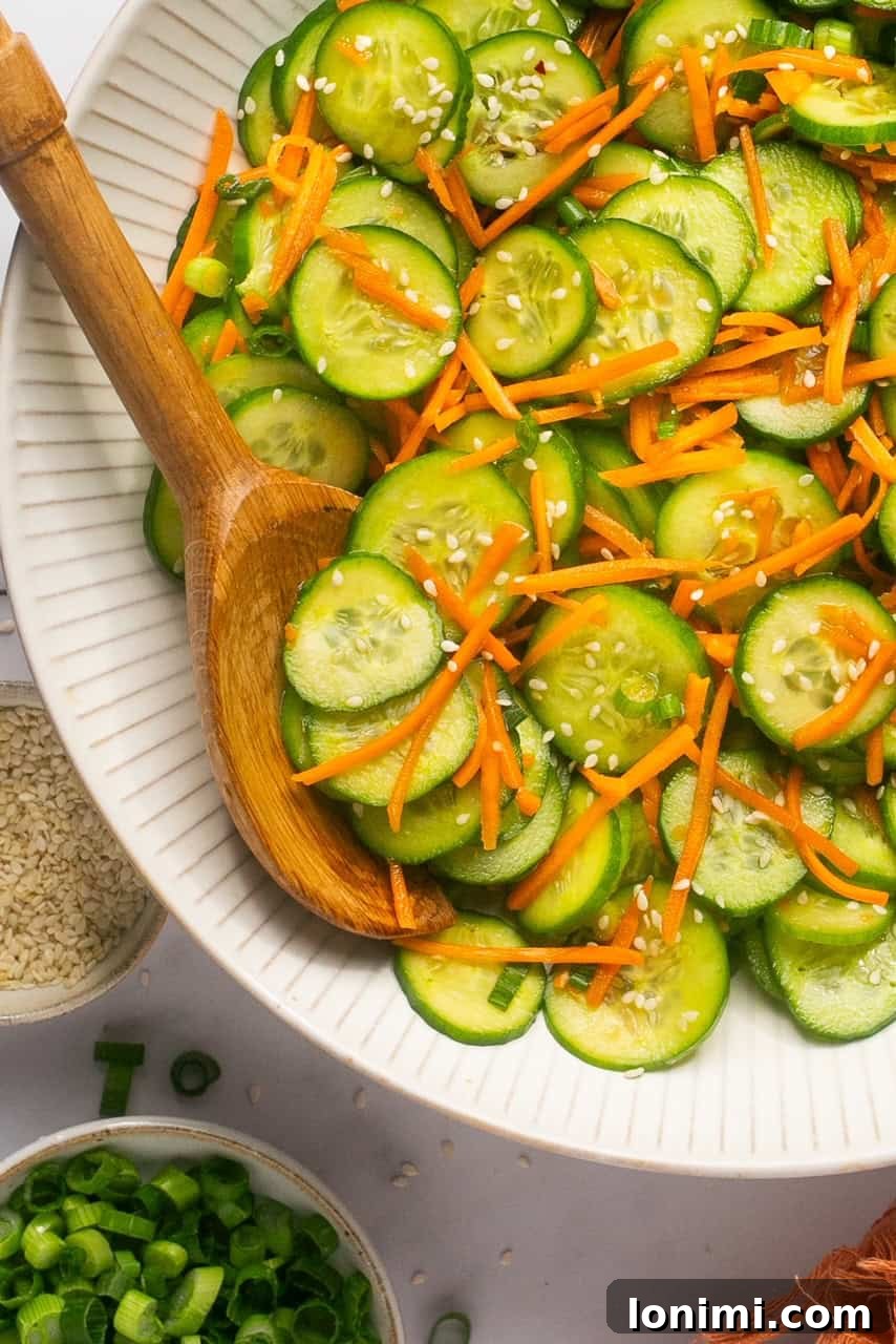 a bowl filled with sliced cucumbers, carrots, and sesame seeds with a wooden spoon resting on the side