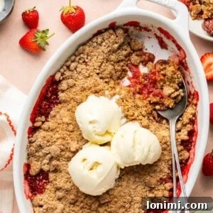 strawberry crumble in an oval baking dish topped with 3 scoops of vanilla ice cream with a spoon taking a portion out