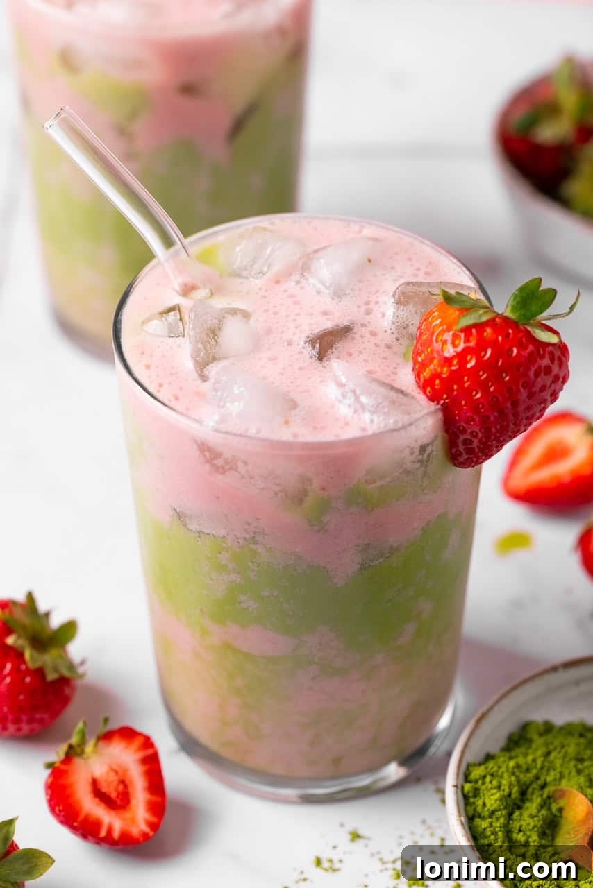 A beautifully layered iced strawberry matcha tea latte in a clear glass, garnished with a fresh strawberry, showcasing vibrant green matcha and soft pink cold foam.