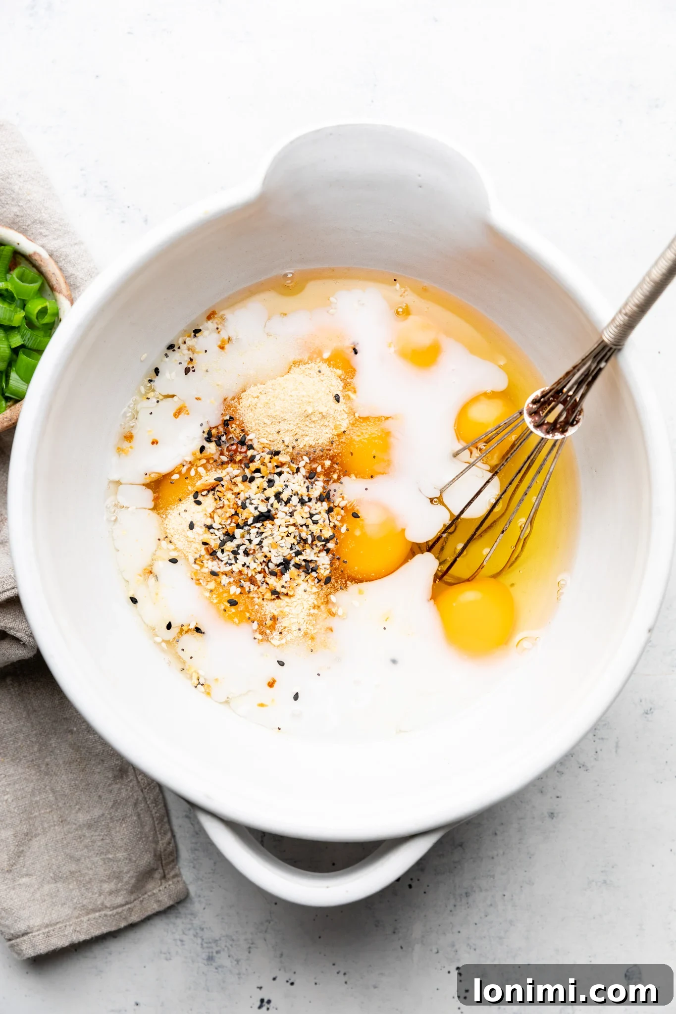 Eggs, dairy-free milk, and everything bagel seasoning ready to whisk in a white mixing bowl on a light colored surface.