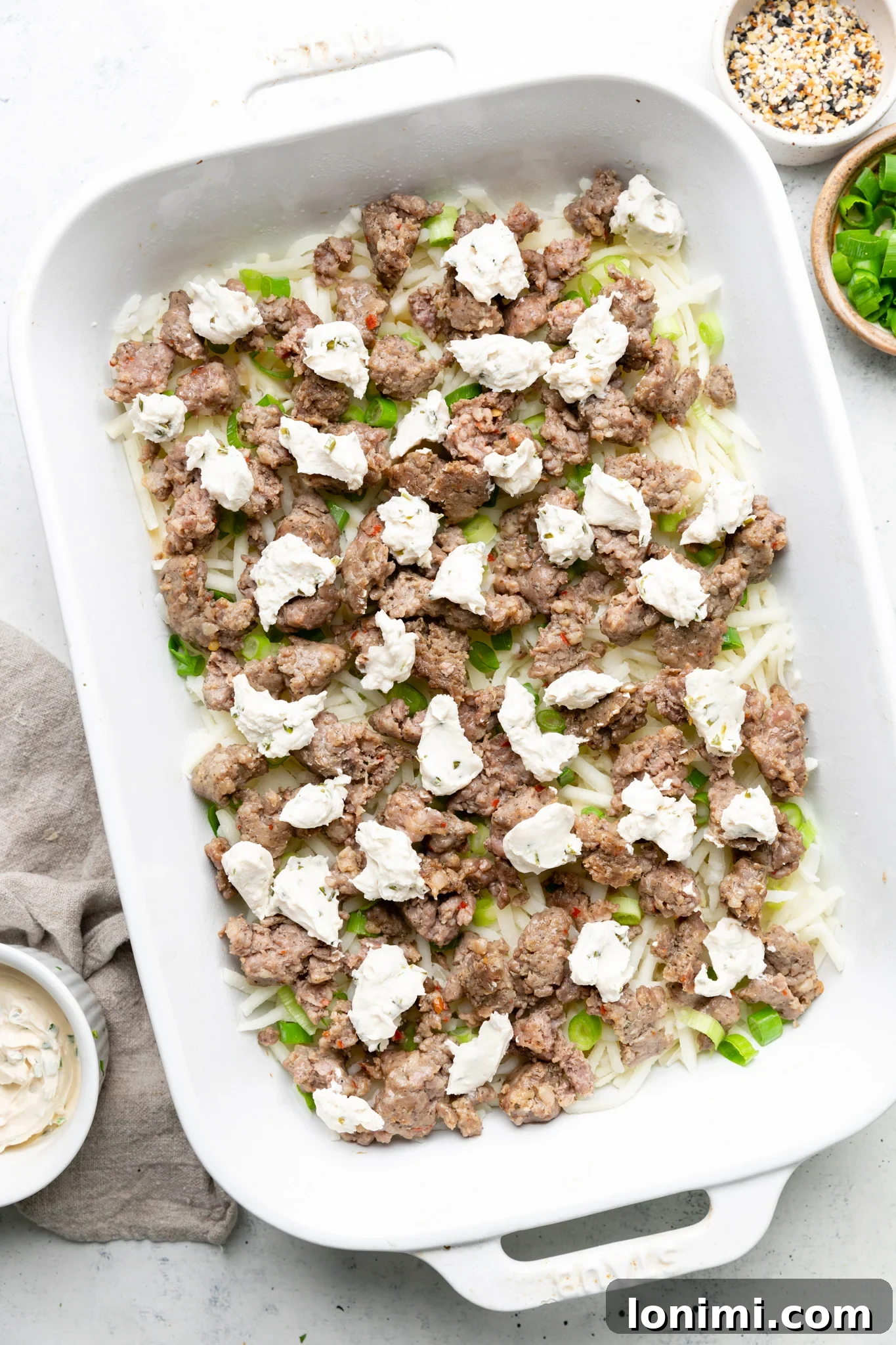 Hash browns, cooked sausage, green onions, and dollops of dairy-free cream cheese layered in a white casserole dish.