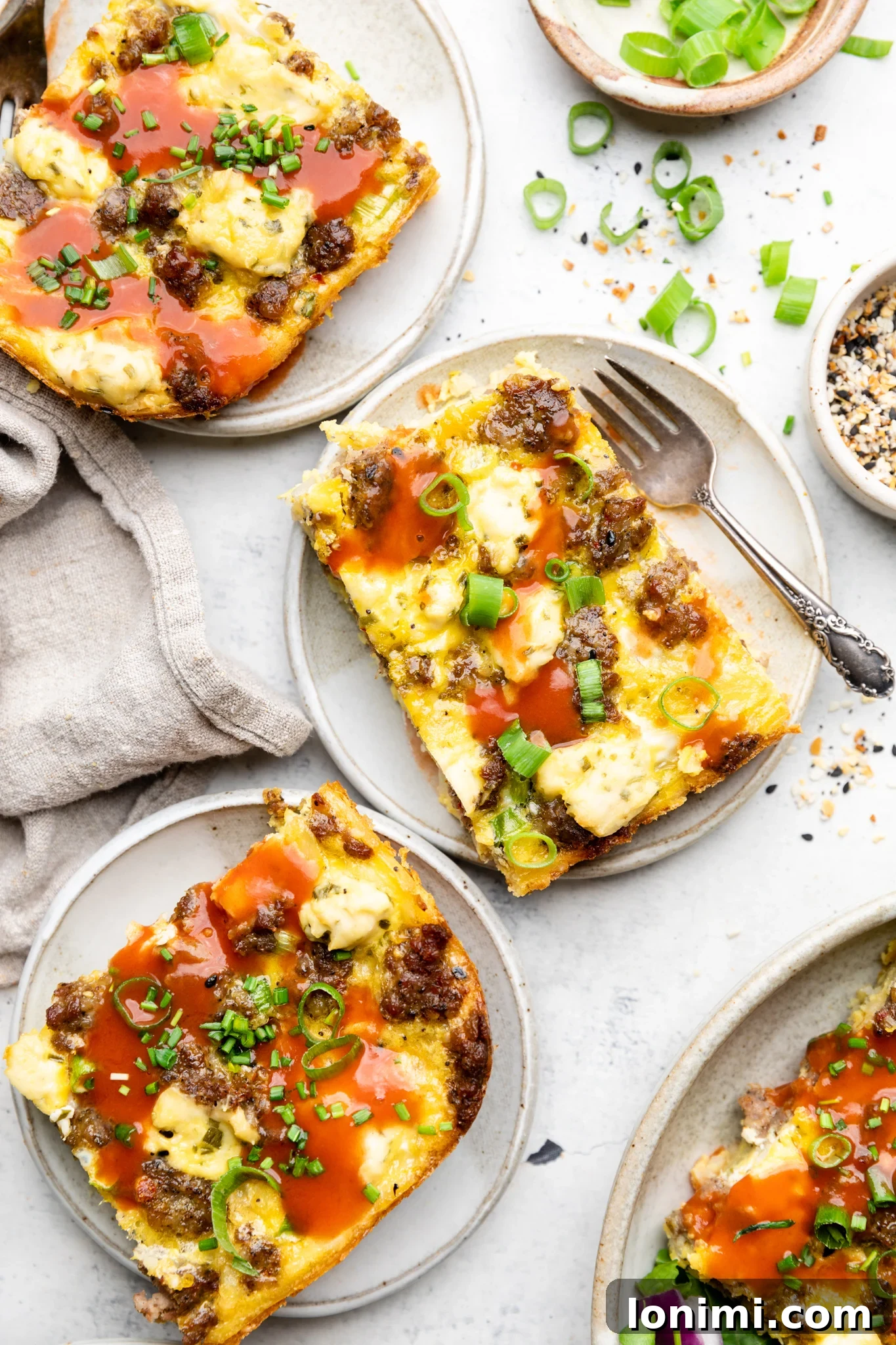 Three slices of sausage hash brown breakfast casserole served on plates with hot sauce and fresh green onions.