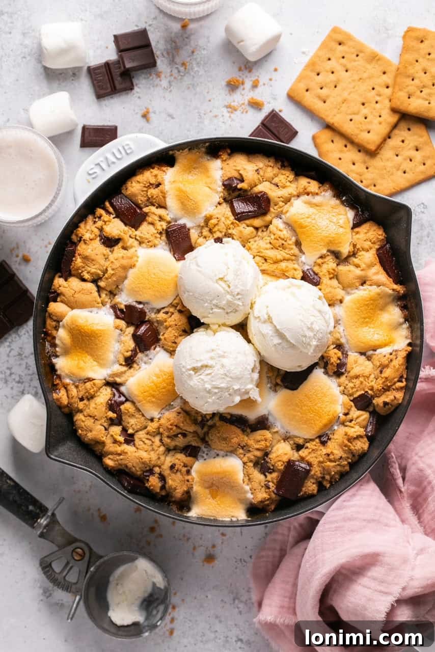 An overhead shot of a chocolate and marshmallow cookie skillet in a cast iron pan topped with vanilla ice cream, with a spoon digging into the gooey center.