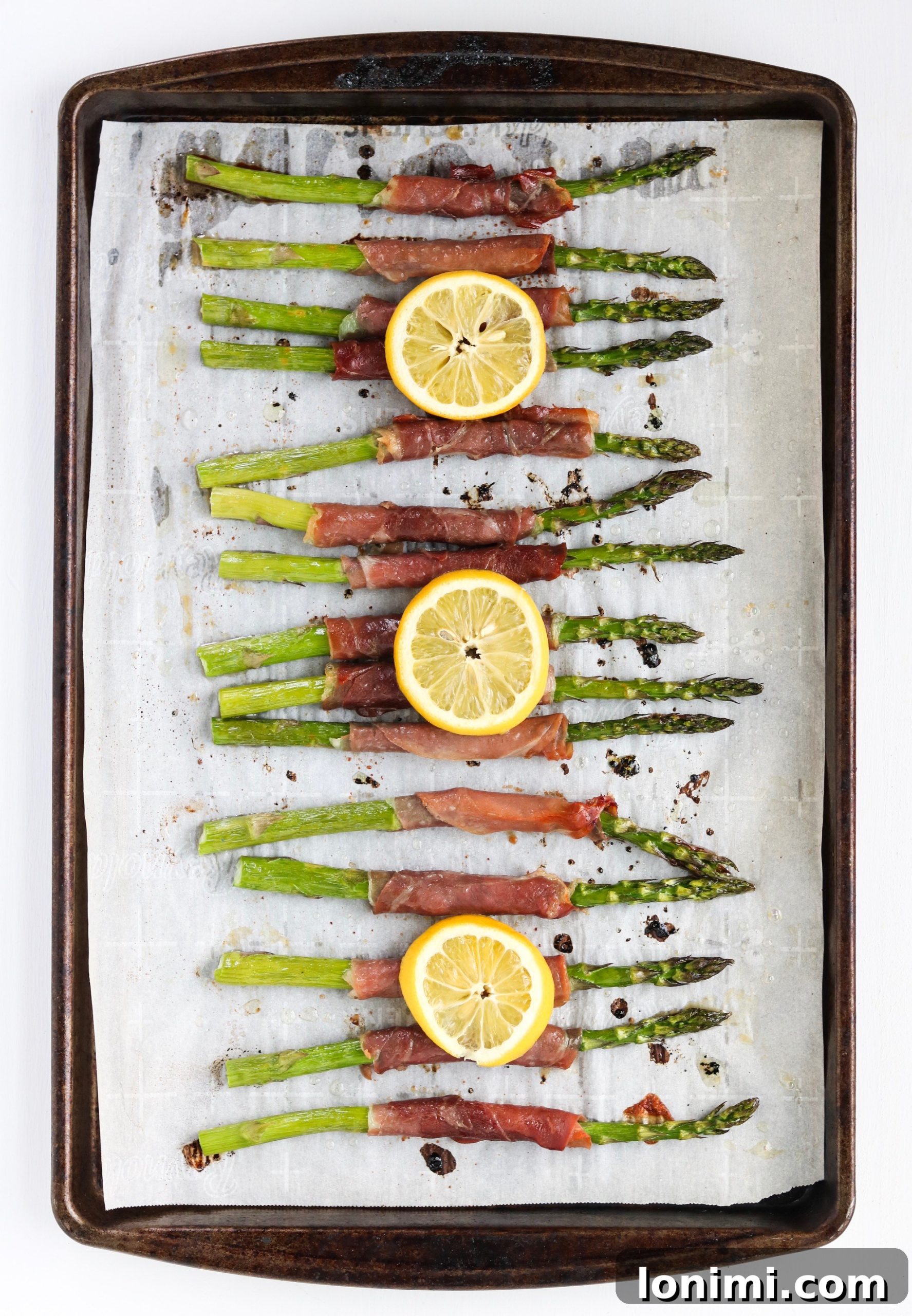 A close-up of a hand wrapping a slice of prosciutto around a fresh green asparagus spear, demonstrating the simple preparation.