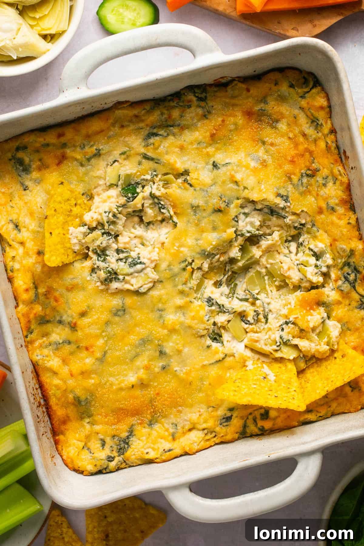 Decadent Mayo-Free Spinach Artichoke Dip 2 Spinach Artichoke Dip in a white square baking dish with two chips dipped in, surrounded by fresh vegetables