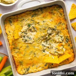 spinach artichoke dip in a white square baking dish with 2 chips dipped in surrounded by vegetables