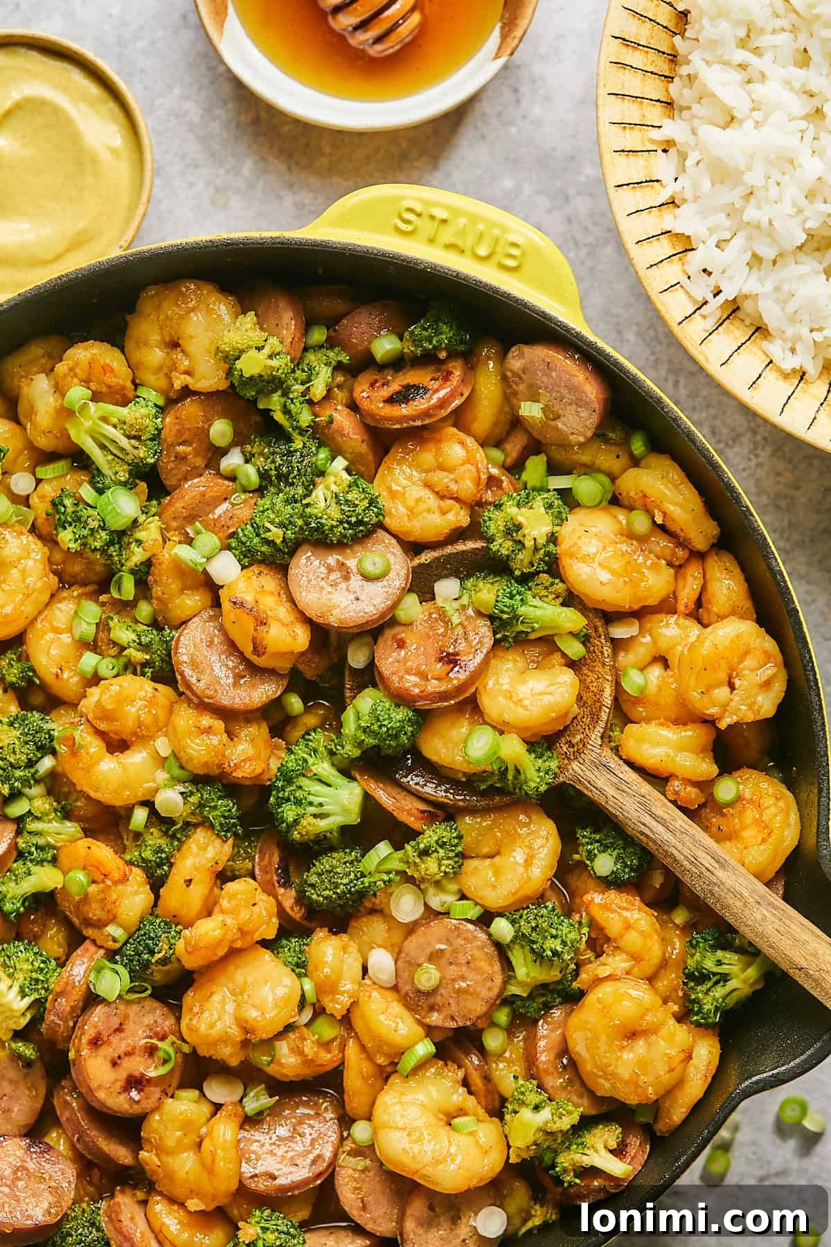 Gluten Free Hot Honey Glazed Shrimp and Sausage Stir Fry 12 A close-up of the delicious hot honey shrimp and sausage stir fry with broccoli, glistening with sauce, in a yellow Staub skillet with a wooden spoon for serving.