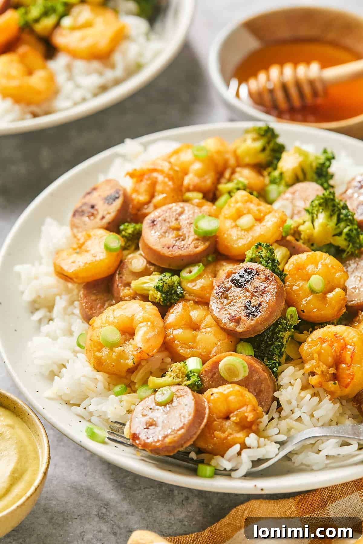 Gluten Free Hot Honey Glazed Shrimp and Sausage Stir Fry 14 A plate of hot honey shrimp and sausage stir fry with broccoli over white rice, with a bowl of honey and chopsticks in the background, showcasing a delightful meal.