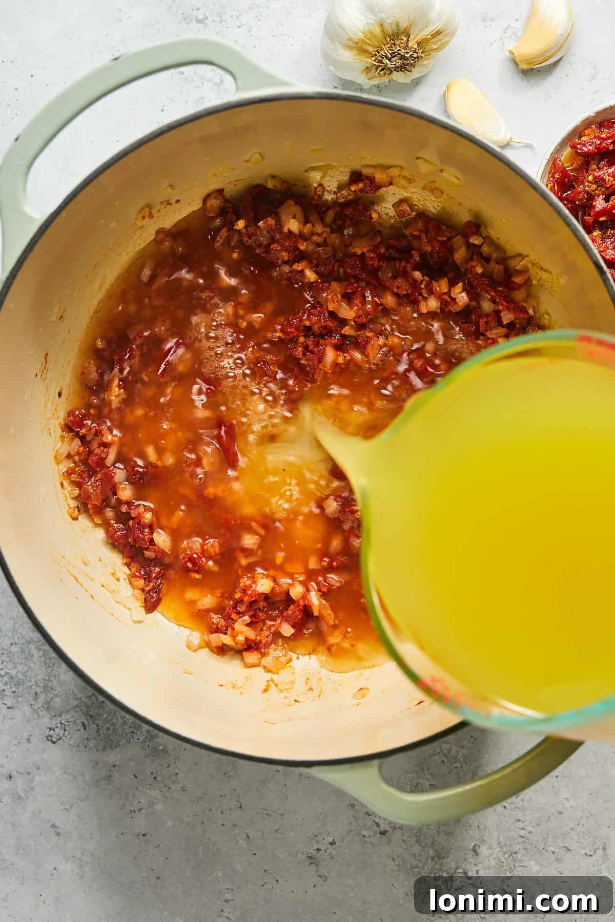 A stream of golden chicken broth being poured into a pot, mixing with the sautéed aromatics and tomato base.