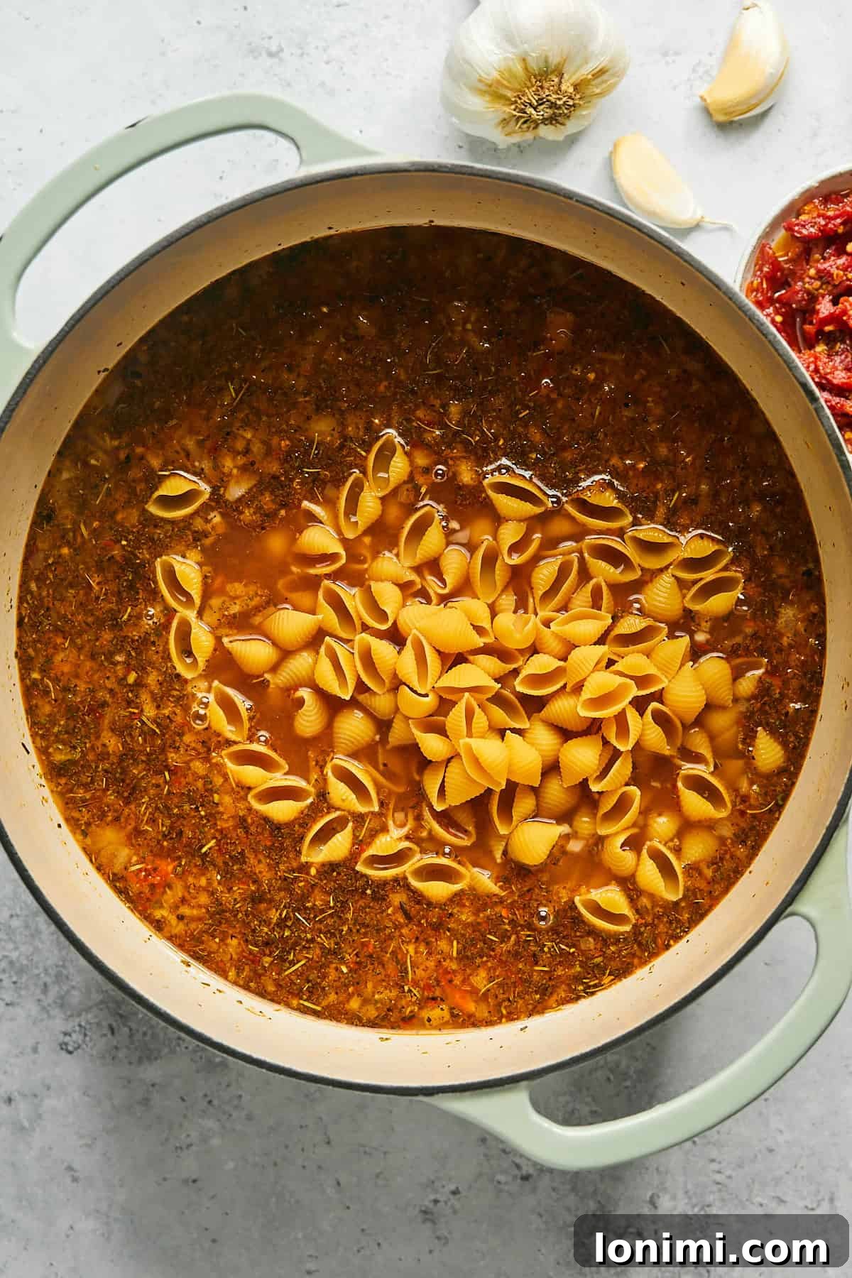 Uncooked gluten-free shell pasta submerged in a rich, simmering red broth inside a large pot.