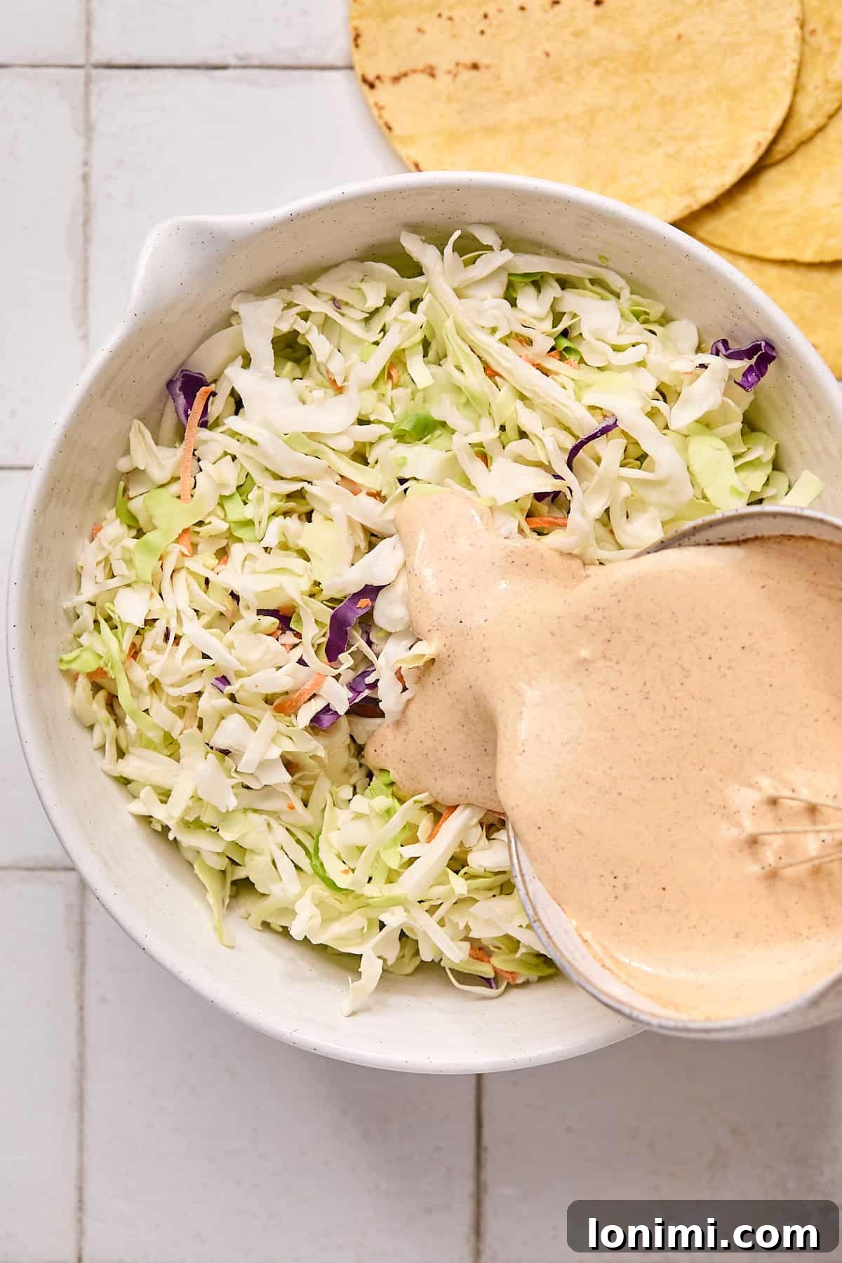 A colorful coleslaw mix in a bowl, with a generous amount of creamy Baja sauce being poured over it, ready to be tossed.