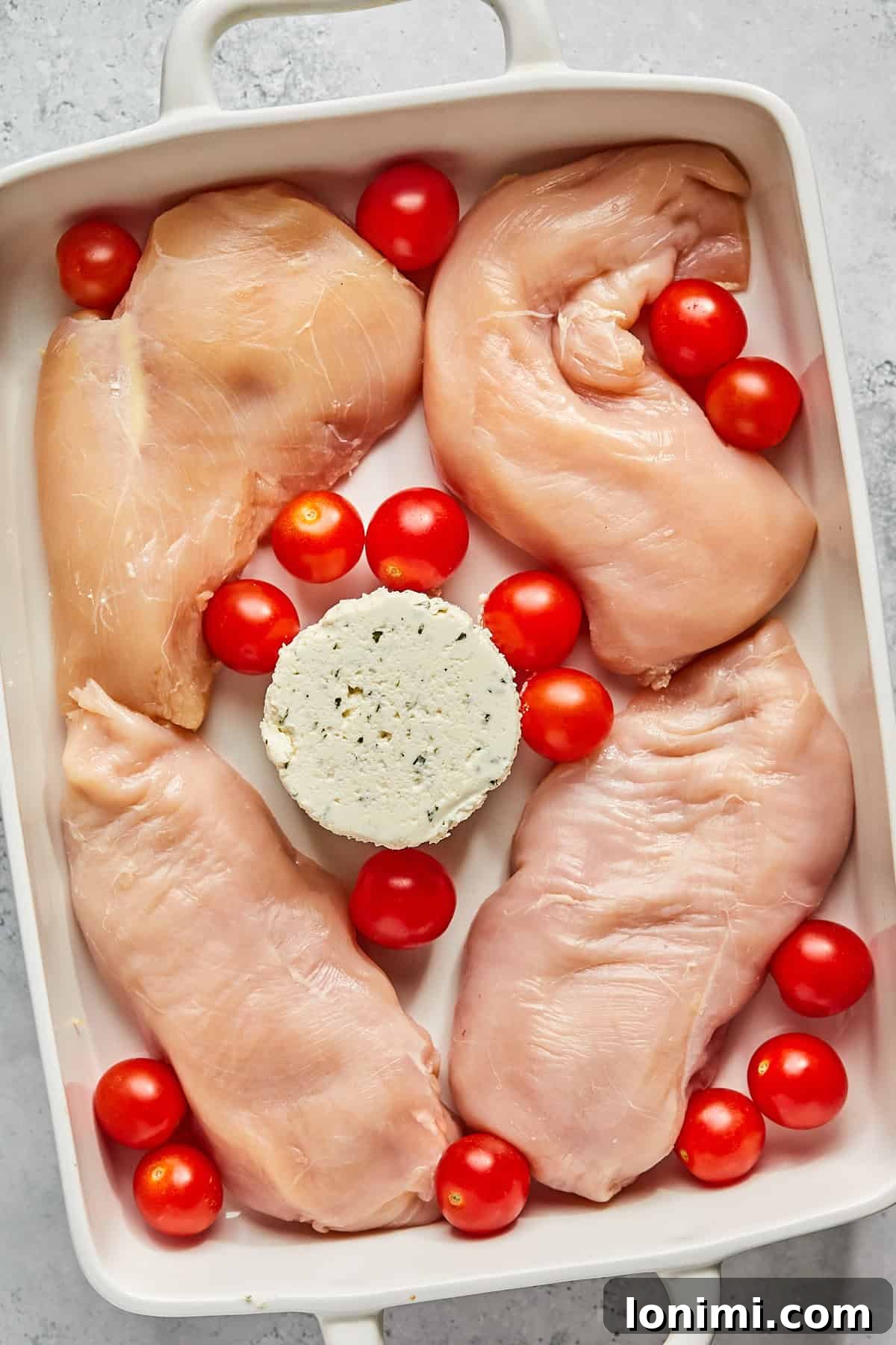 Chicken breasts, cherry tomatoes, and a block of Boursin cheese arranged neatly in a 9x13 inch casserole dish before baking.
