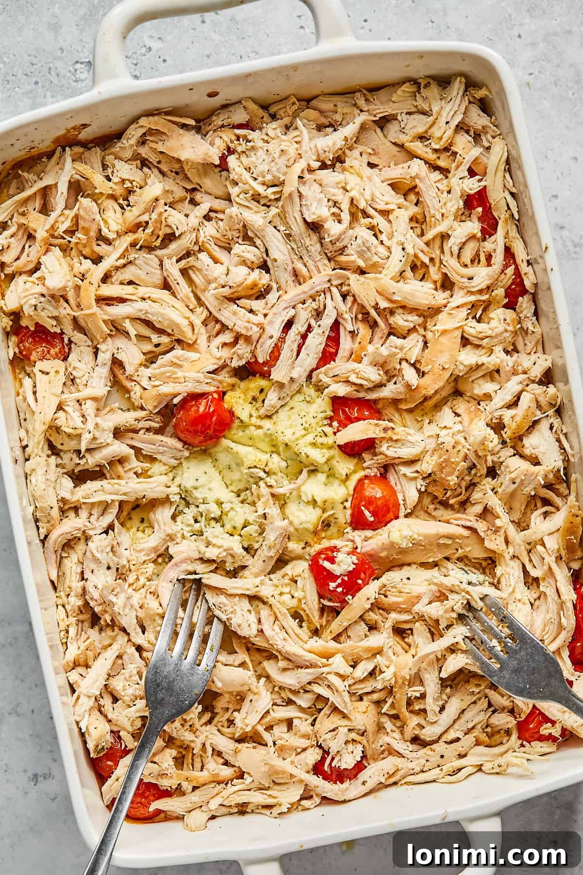 Two forks shredding the perfectly cooked chicken breasts directly in the casserole dish, amidst the melted Boursin and tomatoes.