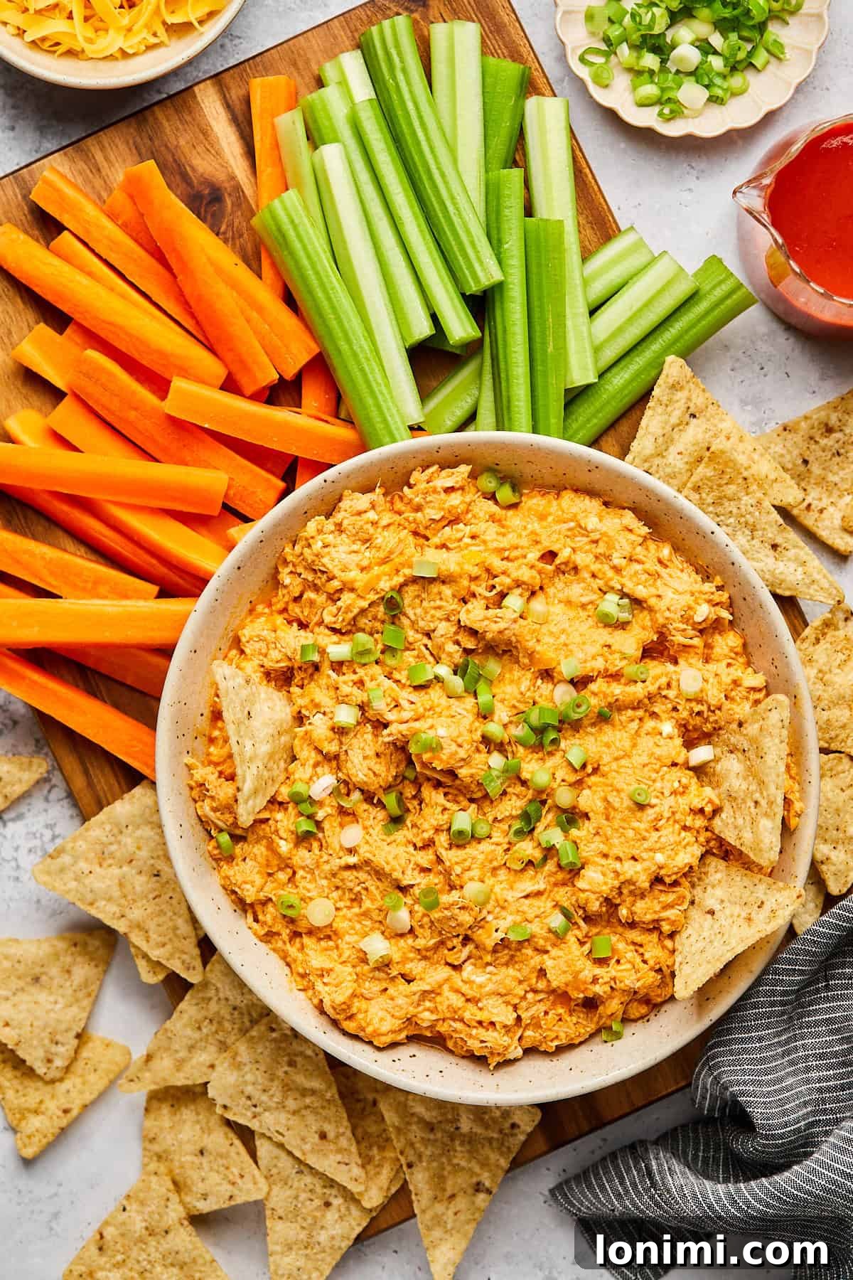 A beautifully presented bowl of high protein buffalo chicken dip, surrounded by a vibrant assortment of tortilla chips and fresh vegetables for dipping.