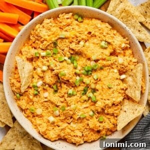 A white bowl filled with high protein buffalo chicken dip, garnished with green onions and surrounded by an array of chips and vegetables for dipping.