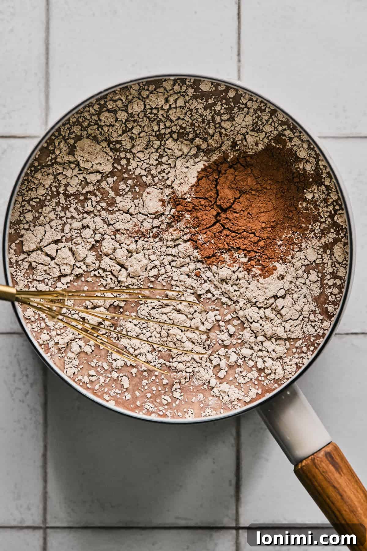 A saucepan on a stove with milk, chocolate protein powder, and cacao powder being whisked together, showing the initial stages of making protein hot chocolate.