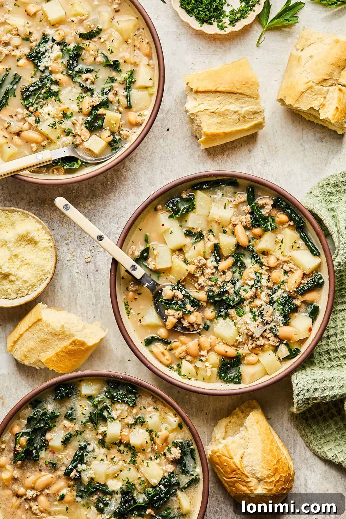 Three bowls of hearty Sausage Kale and White Bean Soup, garnished with fresh herbs and served alongside crusty slices of baguette, ready for a cozy meal.