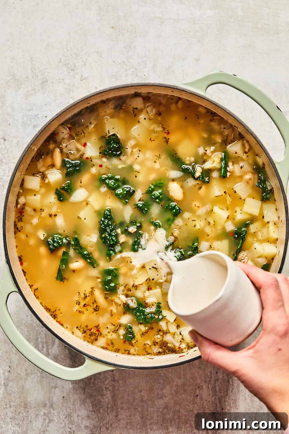 A hand pouring half & half into a pot of creamy Sausage Kale and White Bean Soup.