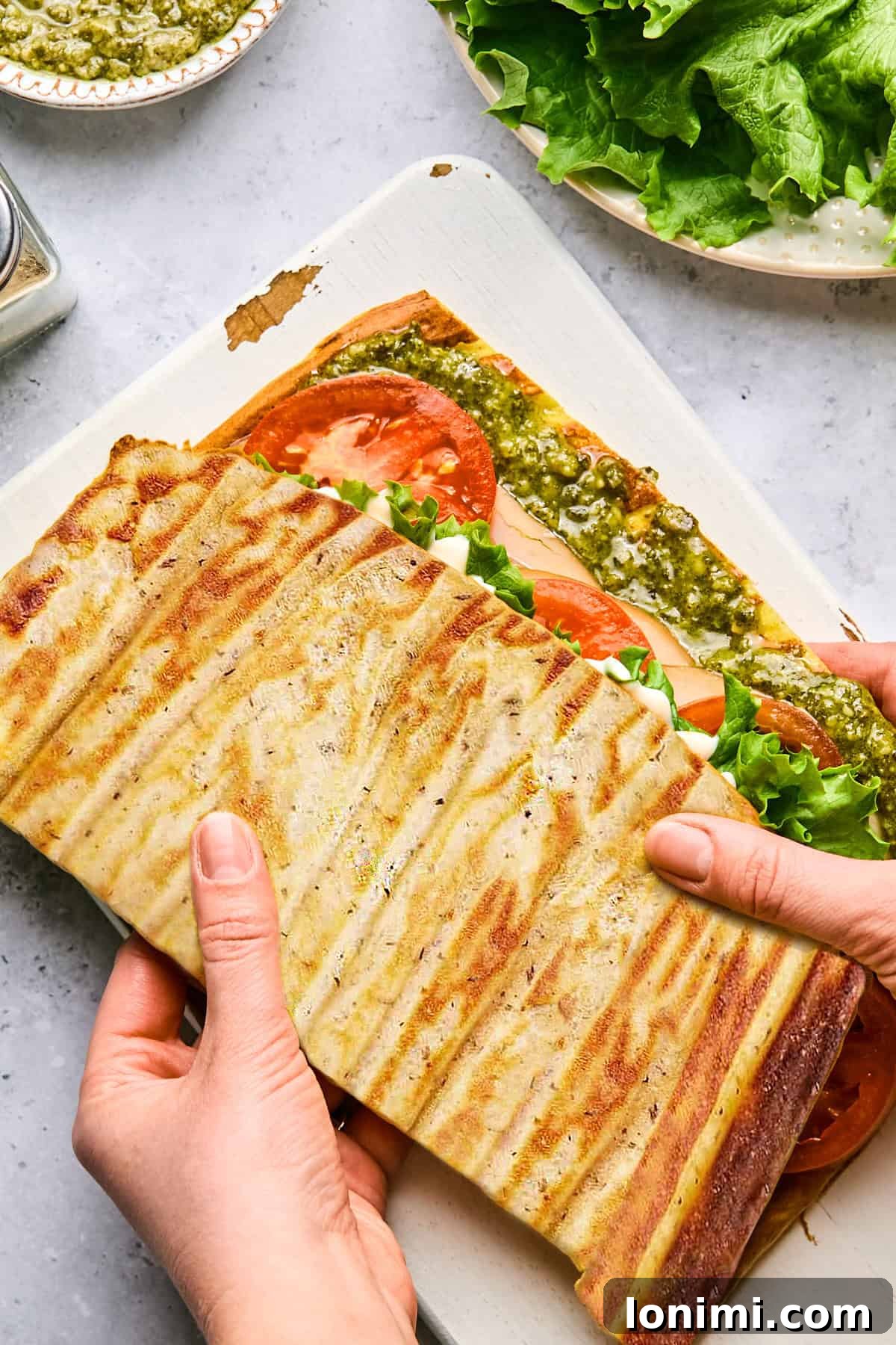 cottage cheese flatbread with turkey, tomato, lettuce and pesto with 2 hands holding it on a cutting board
