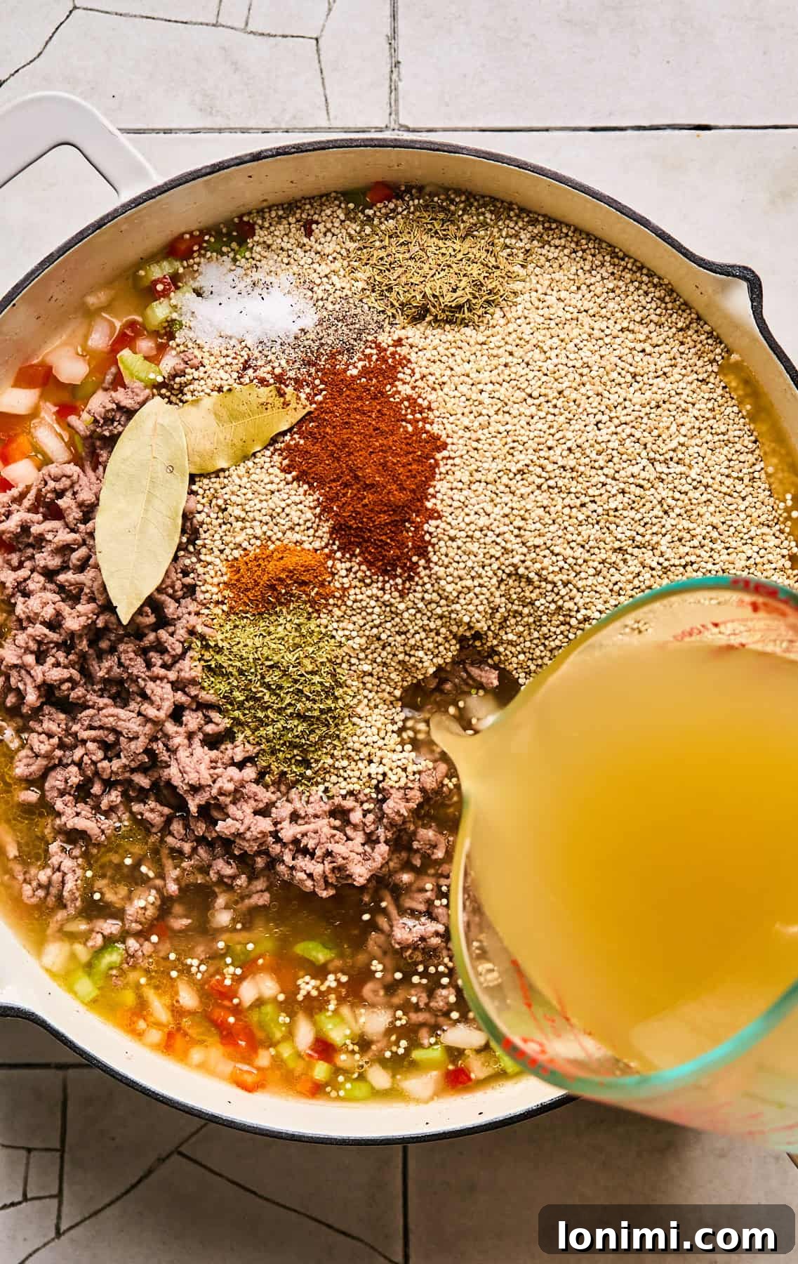 All ingredients – uncooked quinoa, cooked ground beef, chicken broth, bay leaves, and spices – being added to the sautéed vegetables in a skillet.