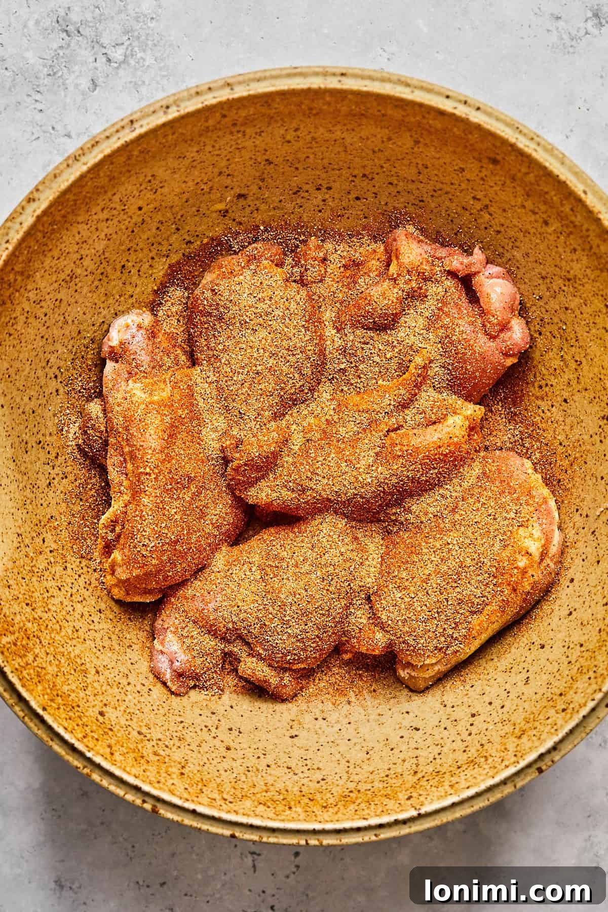 Raw chicken thighs in a mixing bowl are generously coated with a dark, savory spice blend, awaiting their transformation into a delicious meal.