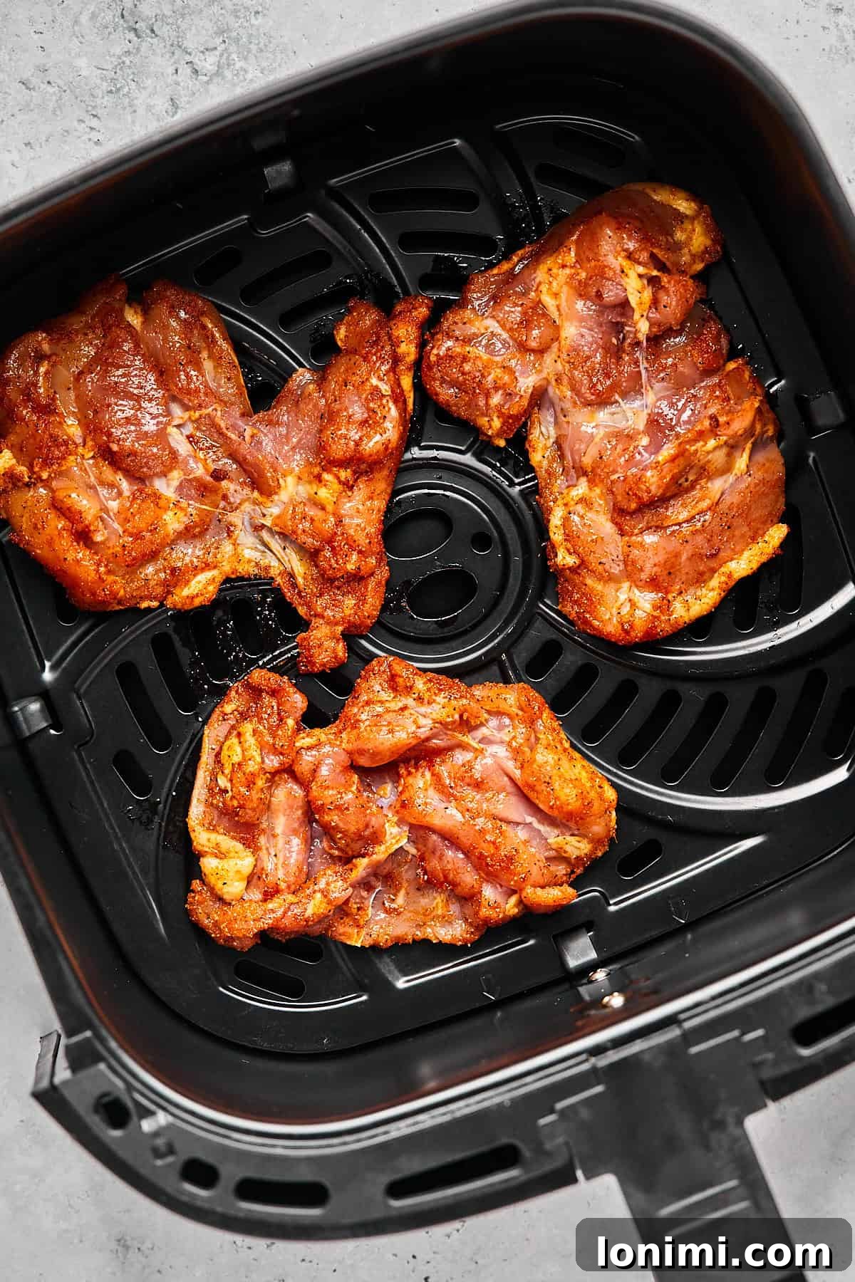 Several raw, spice-coated boneless chicken thighs are meticulously arranged in a single, even layer within the basket of an air fryer, ready for cooking.