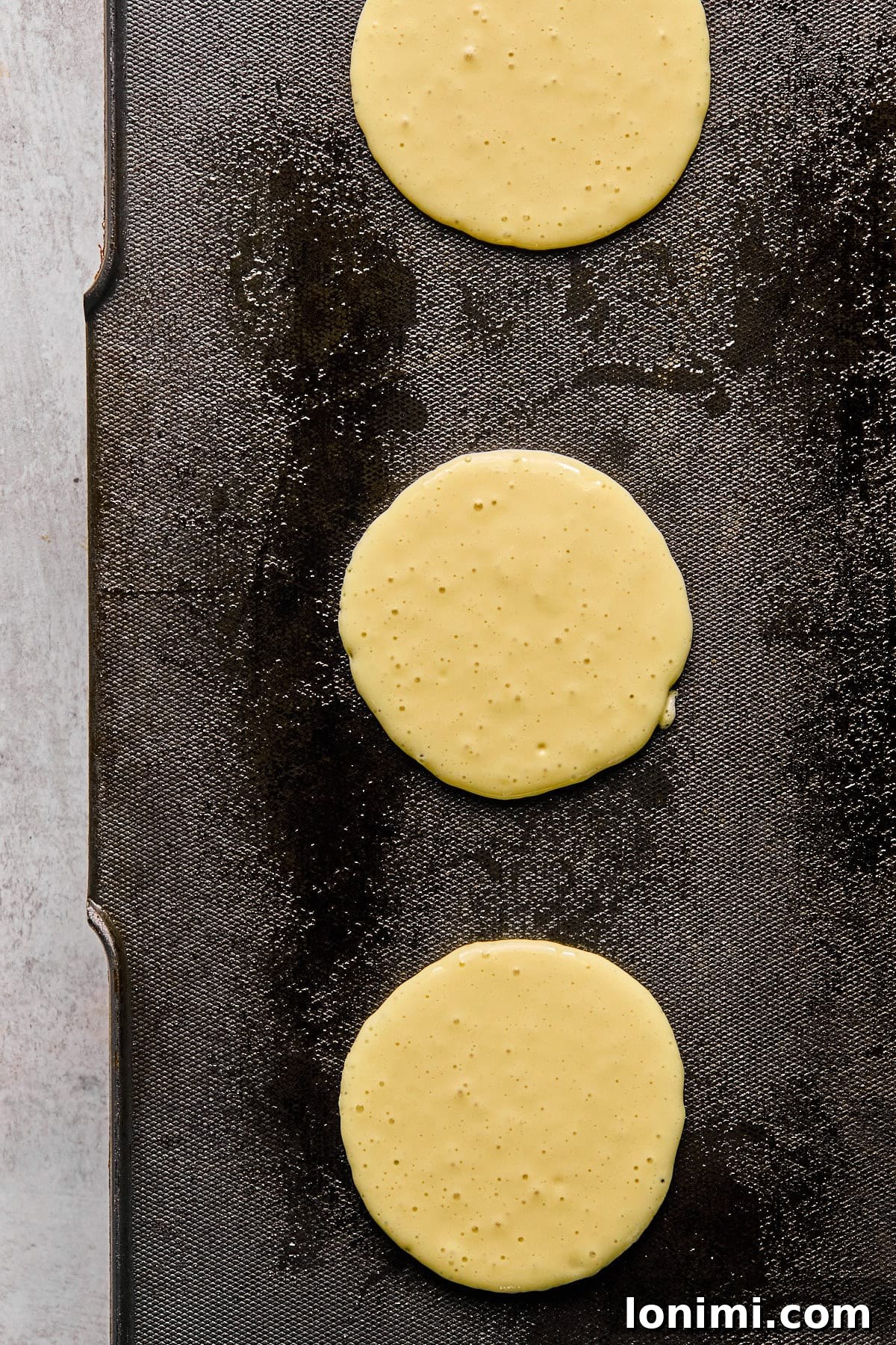Ultimate Cottage Cheese Protein Pancakes 6 Three circles of uncooked pancake batter cooking on a hot griddle, starting to form small bubbles on their surface.