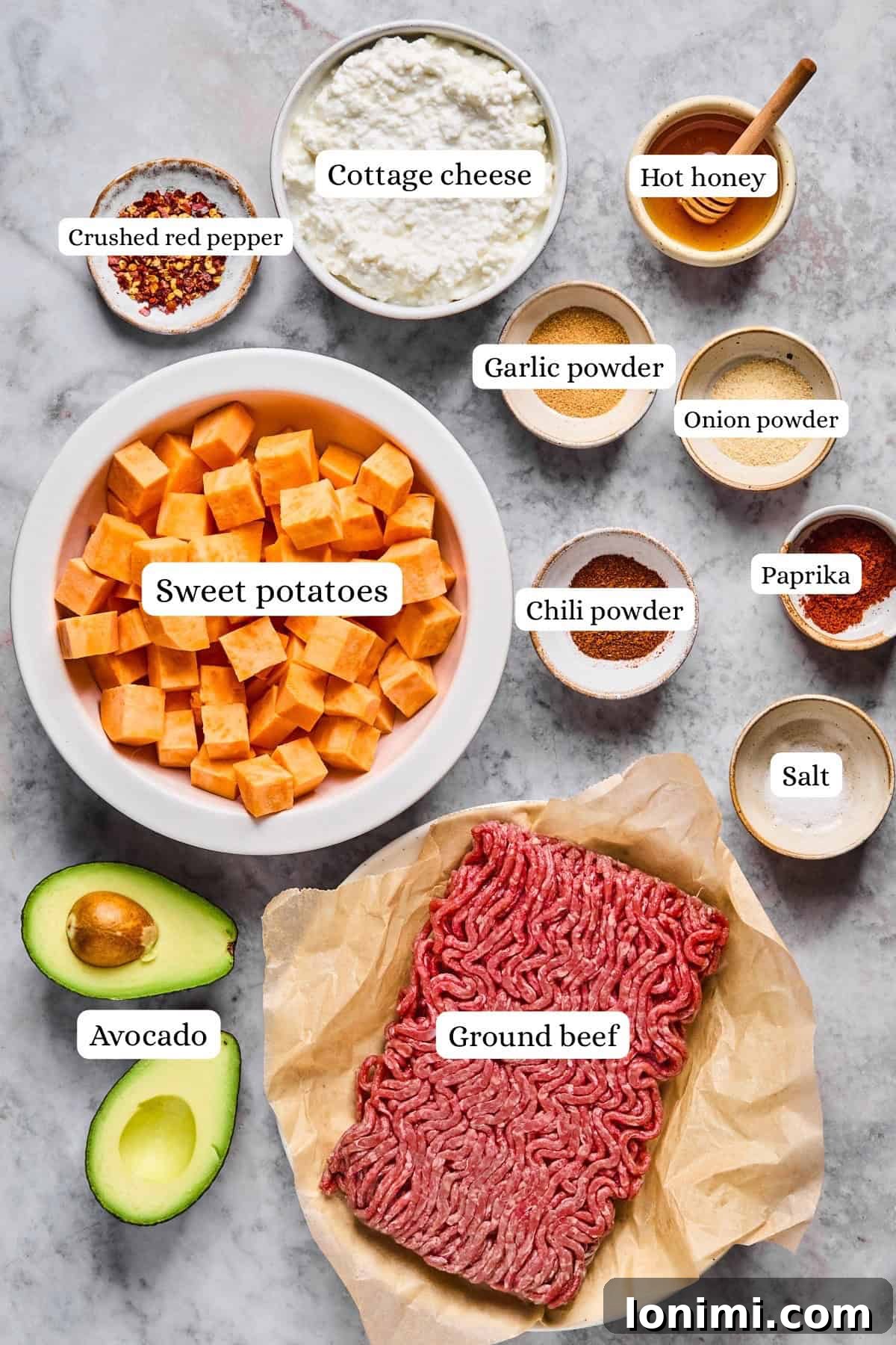 The Trending Protein Power Cottage Cheese Taco Bowl 3 Image showing labeled ingredients on a surface: a bowl of cubed sweet potatoes, a dish of cottage cheese, ground beef, a sliced avocado, and small bowls containing crushed red pepper, hot honey, paprika, salt, chili powder, garlic powder, and onion powder.