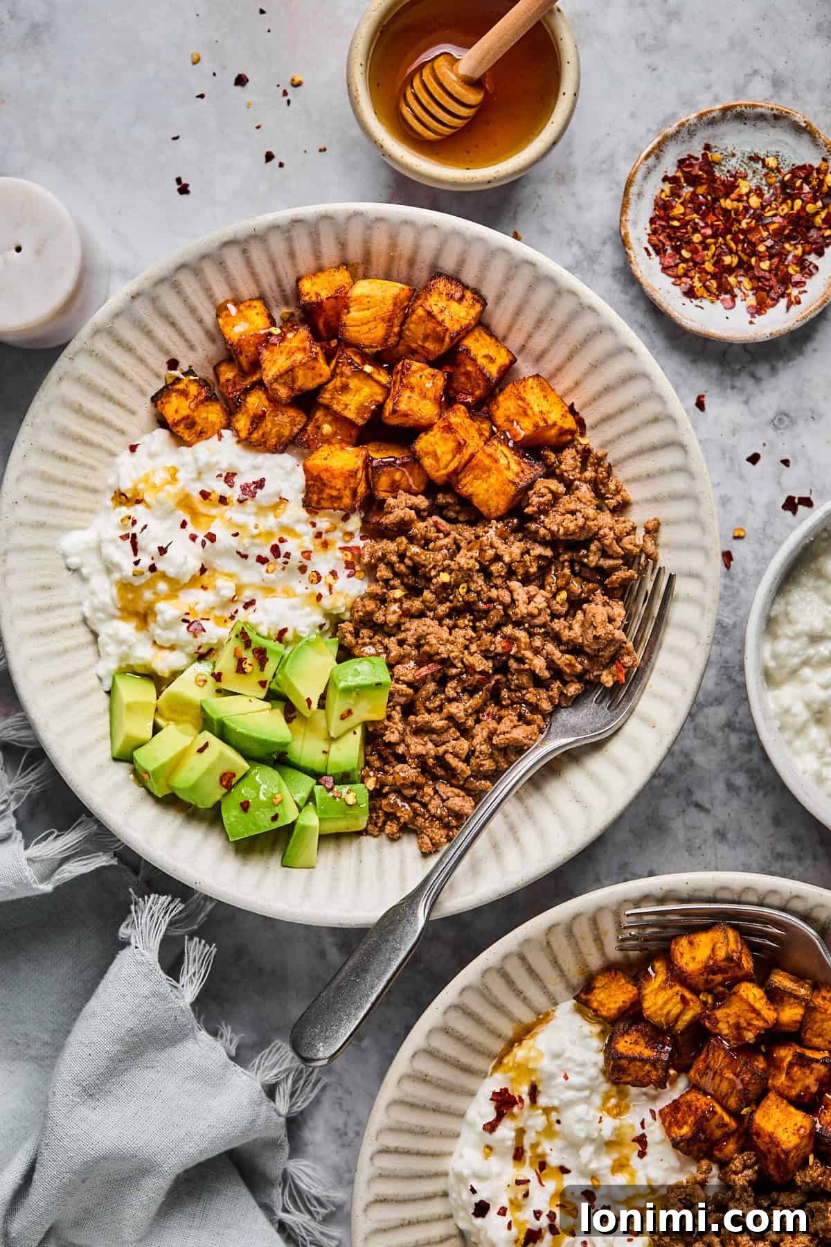 The Trending Protein Power Cottage Cheese Taco Bowl 8 Cottage cheese taco bowl made with seasoned ground meat, roasted sweet potatoes, sliced avocado, and cottage cheese with red pepper flakes. A fork rests on one plate. Honey, red pepper flakes, a napkin, and a small candle nearby.