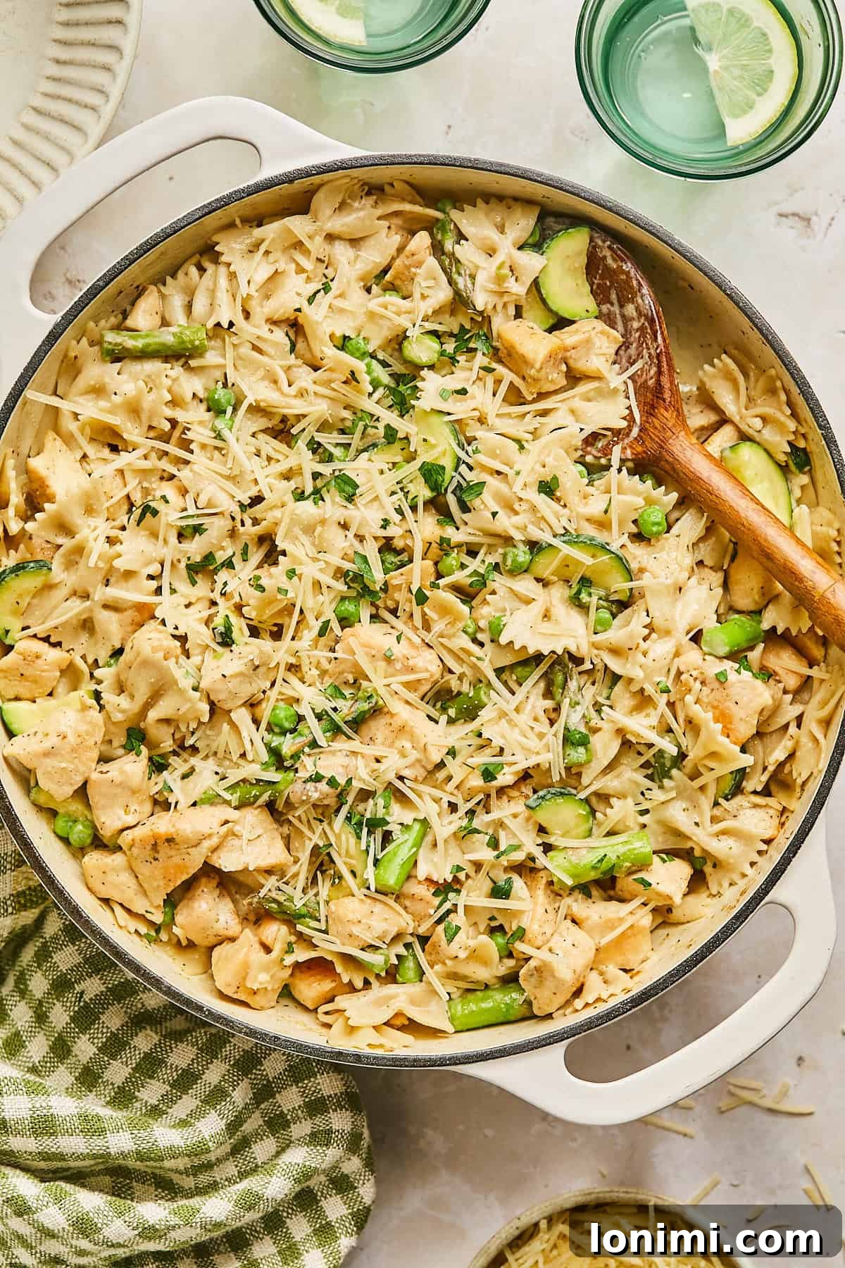 A large pot of creamy pasta featuring bow tie noodles, chicken chunks, asparagus, and zucchini. Topped with grated cheese and herbs, a wooden spoon rests in the pot. Two glasses of water with lemon slices are beside the pot, next to a checkered cloth.