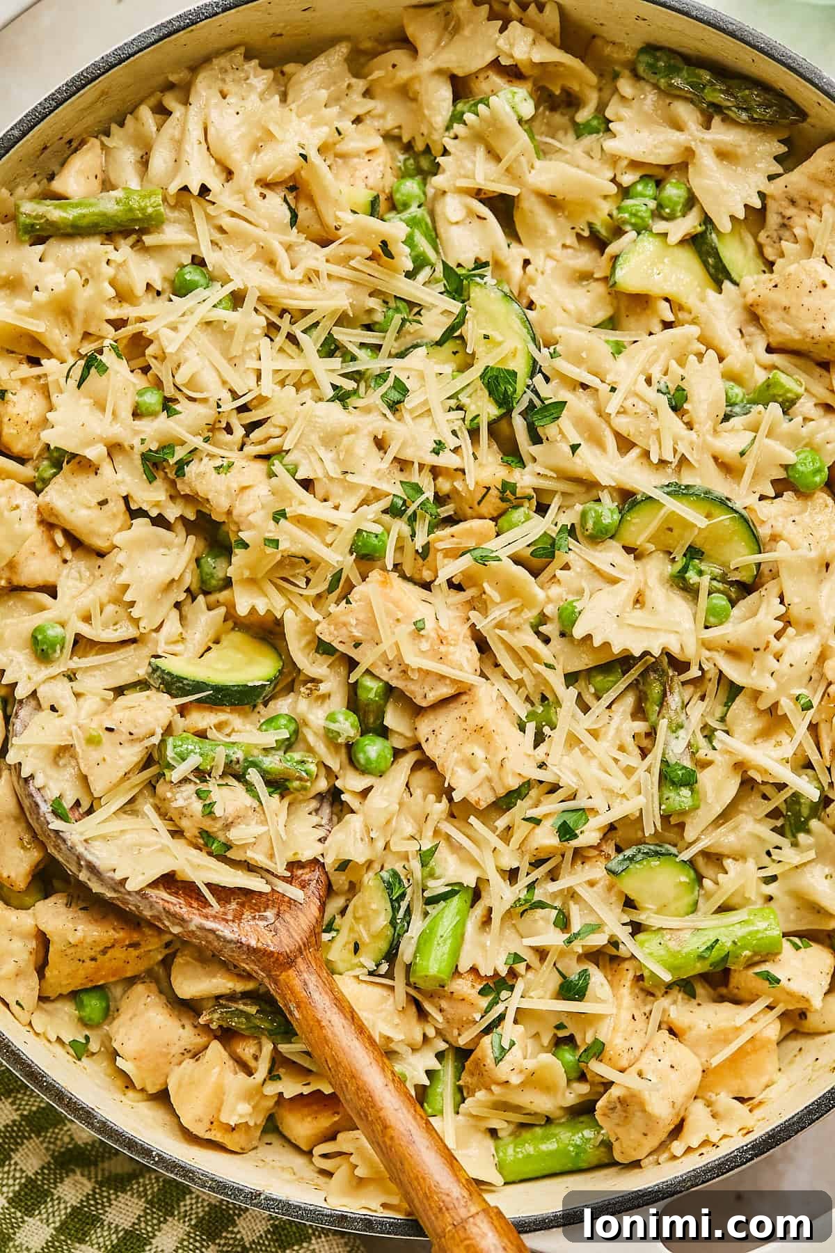 A skillet filled with boursin chicken pasta with bow tie pasta, chunks of chicken, asparagus, zucchini, and peas, topped with shredded Parmesan cheese and garnished with herbs. A wooden spoon rests in the pasta.