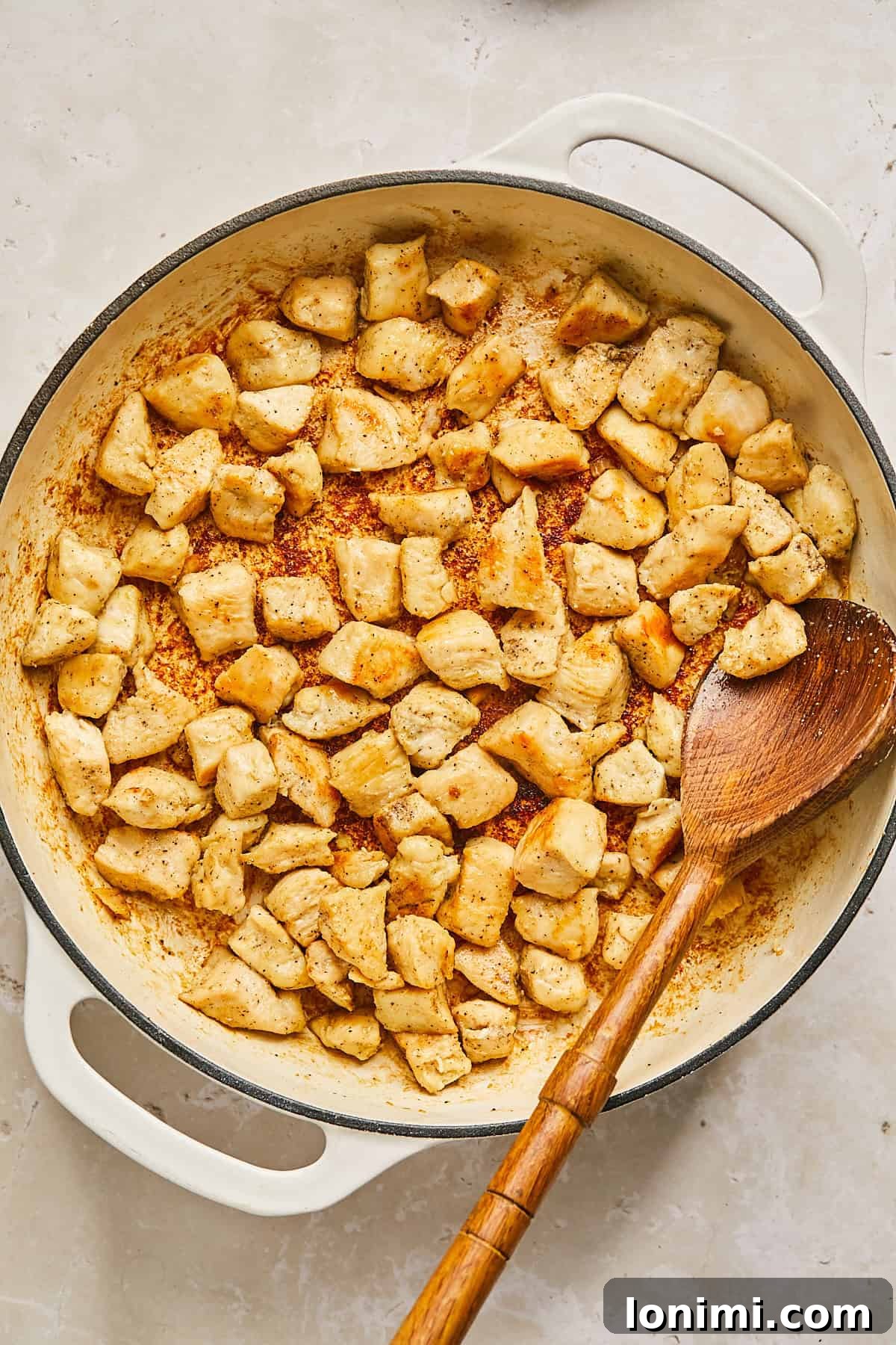 A white skillet filled with cooked, golden-brown chicken pieces, seasoned with spices. A wooden spoon rests in the skillet, suggesting the chicken has been stirred or sautéed. The background is a light-colored countertop.