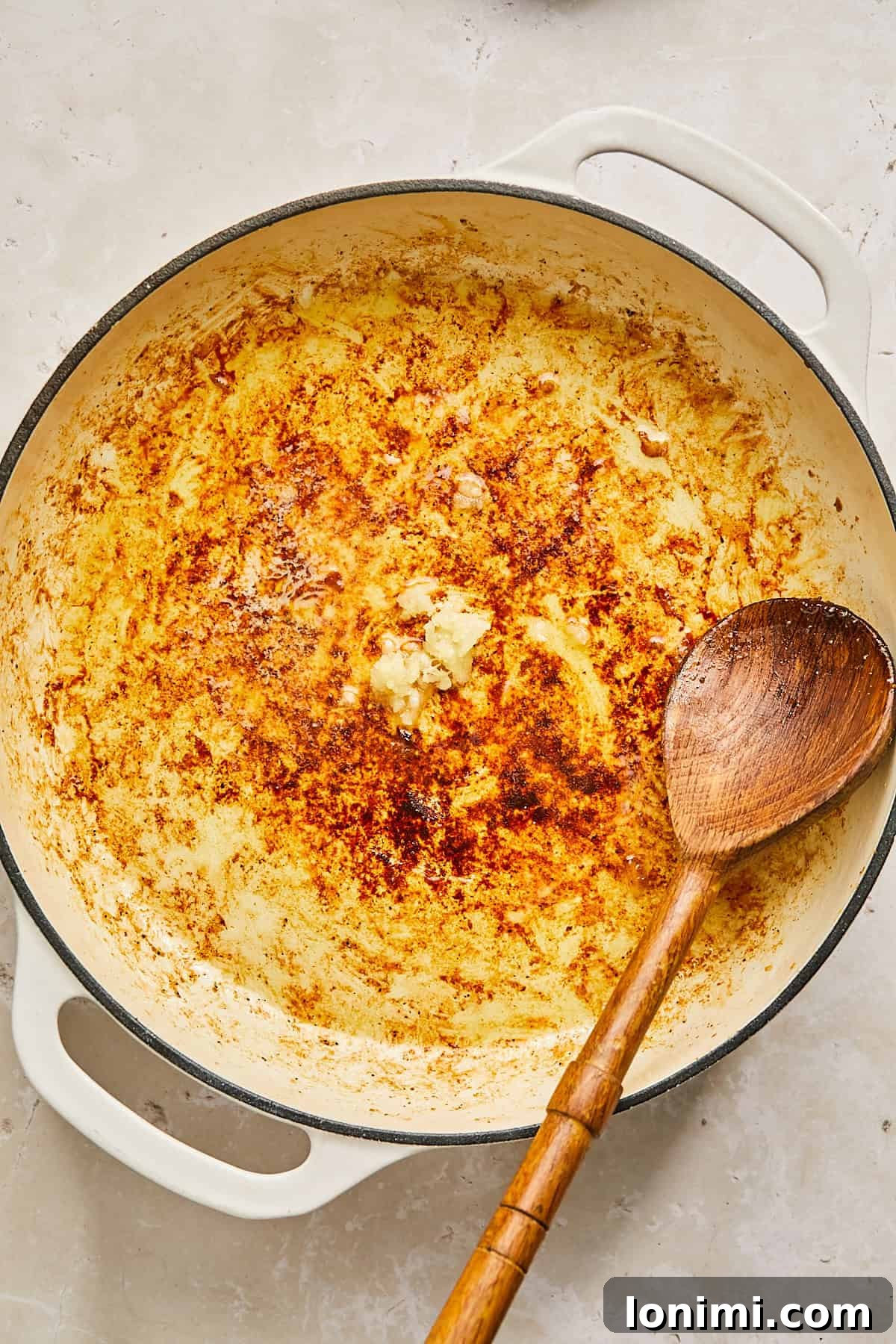 A large, round, white enameled cast iron pan with bits of cooked food and brown residues at the bottom. A wooden spoon rests inside, angled to the right. The background is a light-colored surface.