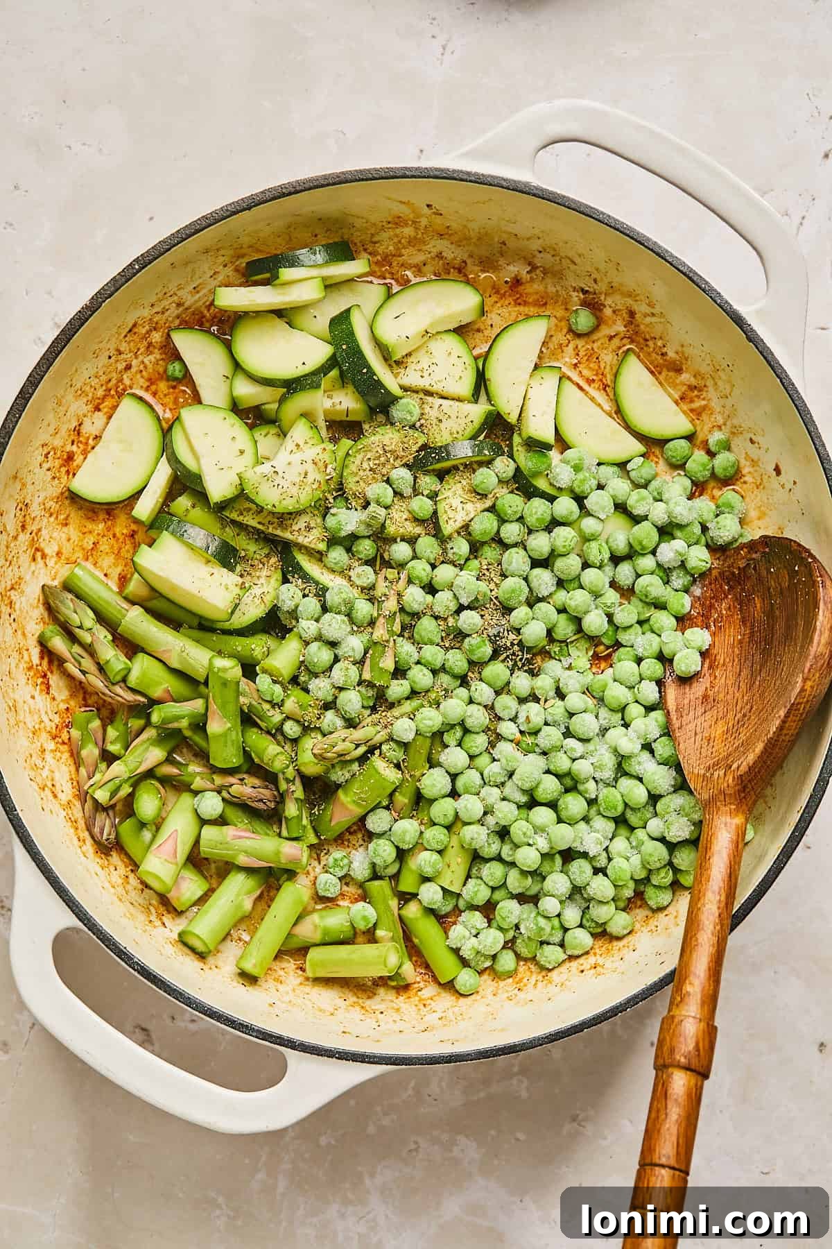 A white pot filled with chopped asparagus, zucchini slices, and frozen peas, placed on a light beige surface. A wooden spoon rests inside the pot, ready to stir the vibrant, fresh ingredients.