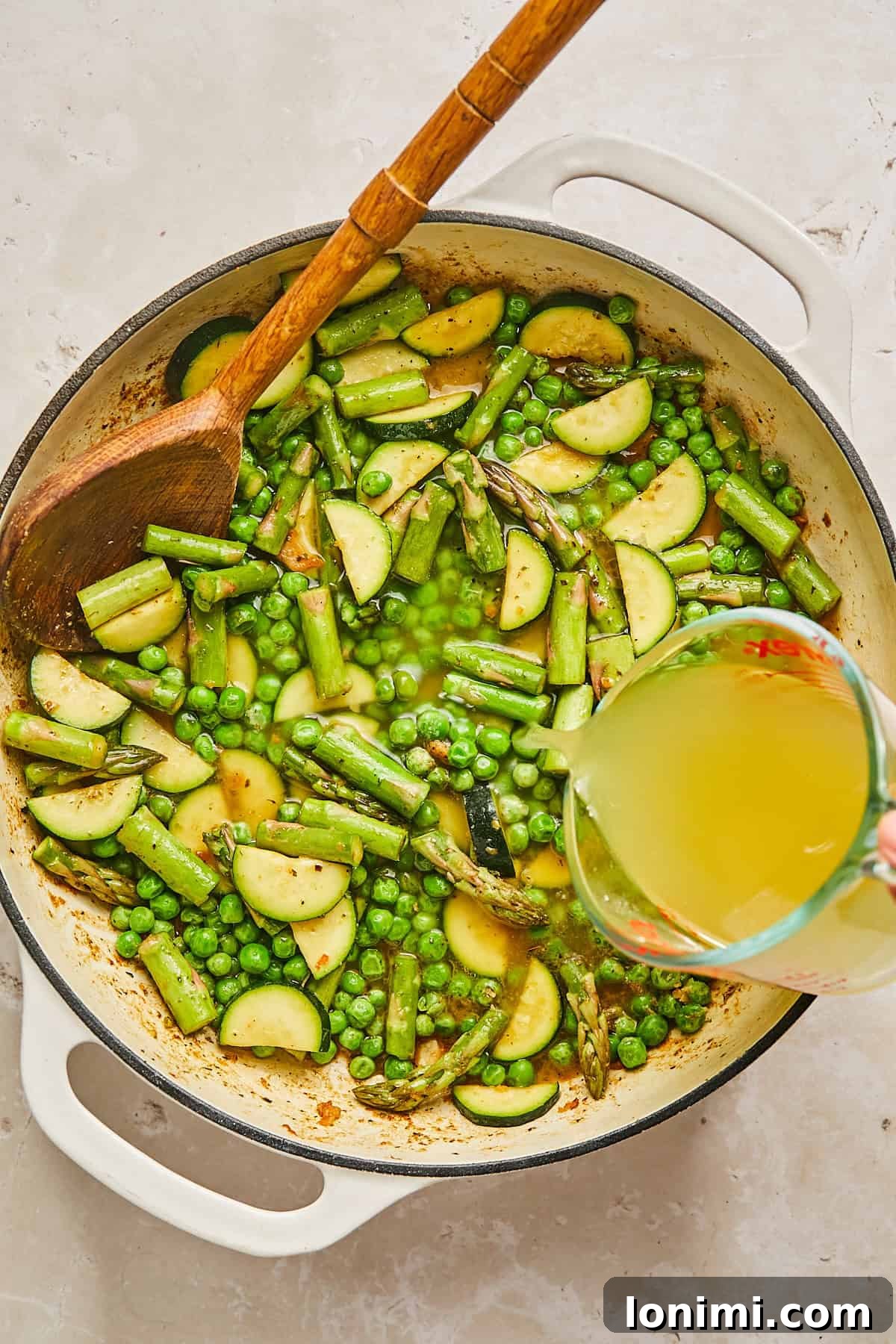 A cast iron pan filled with a mixture of sliced zucchini, asparagus, and peas is placed on a light surface. A wooden spoon rests in the pan, and a measuring cup is pouring broth into the mixture.