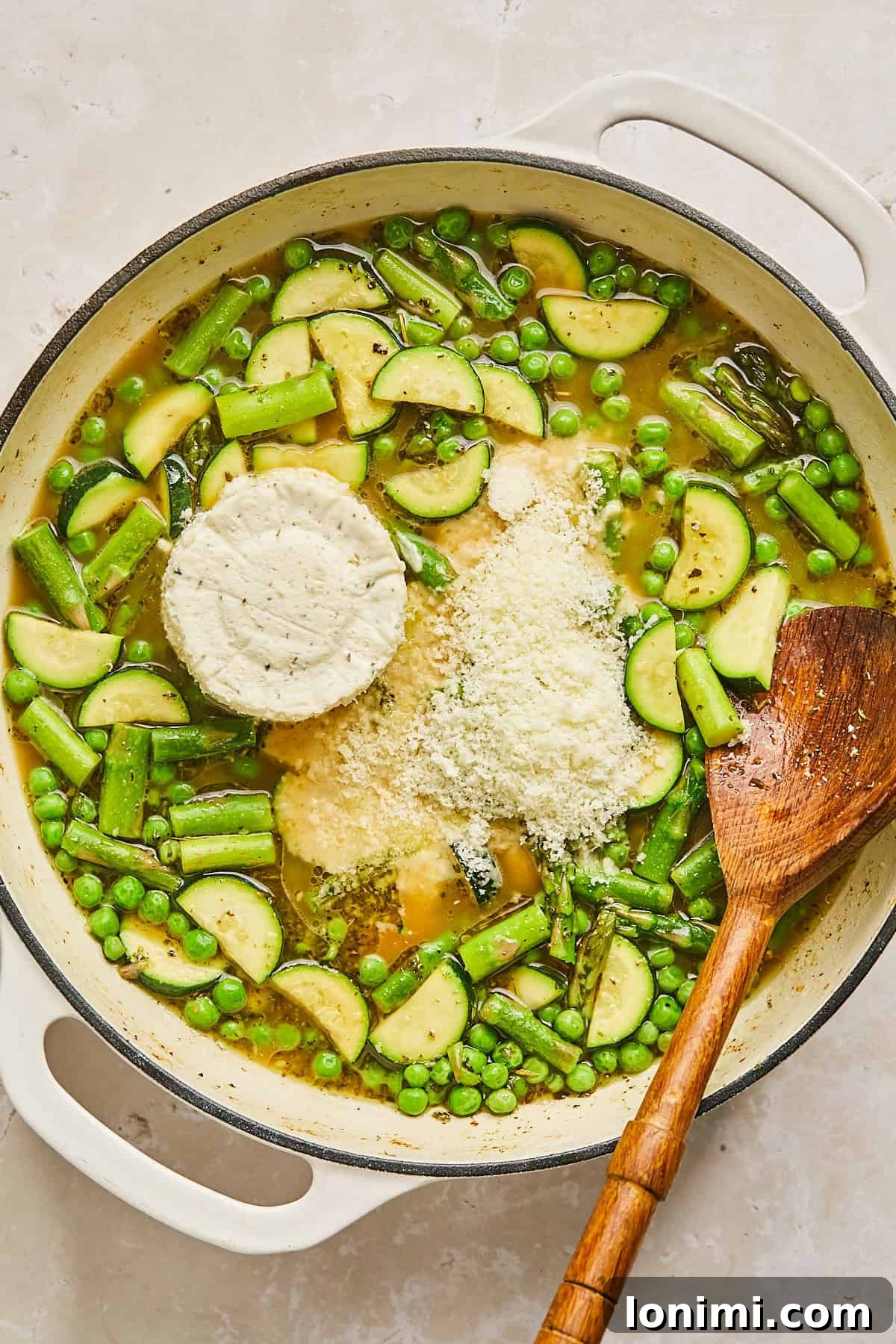 A large white pot filled with a vibrant mixture of zucchini slices, peas, and asparagus in a broth. A round piece of boursin cheese and a heap of grated parmesan are on top. A wooden spoon rests on the right side of the pot.