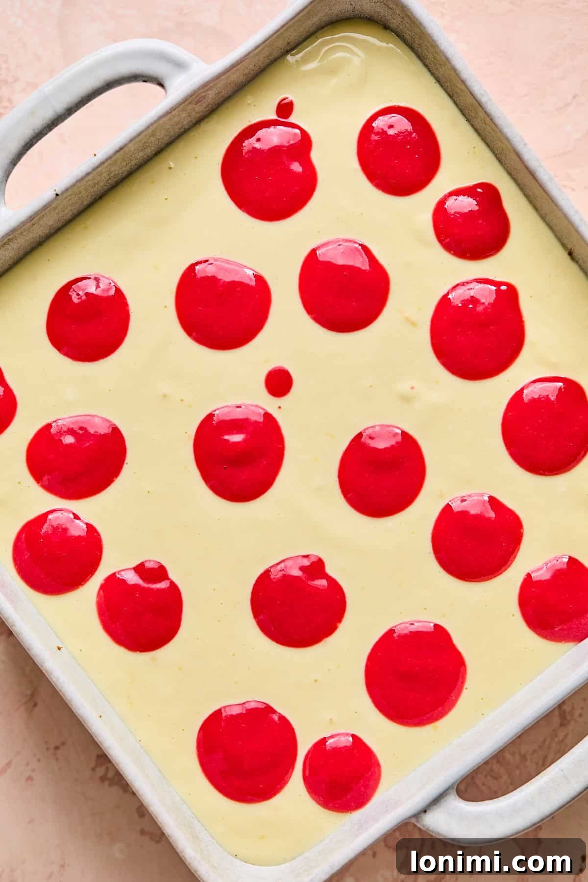 Drops of vibrant raspberry puree artfully spooned onto the smooth, white cheesecake filling mixture in a square pan, preparing for the swirling step.
