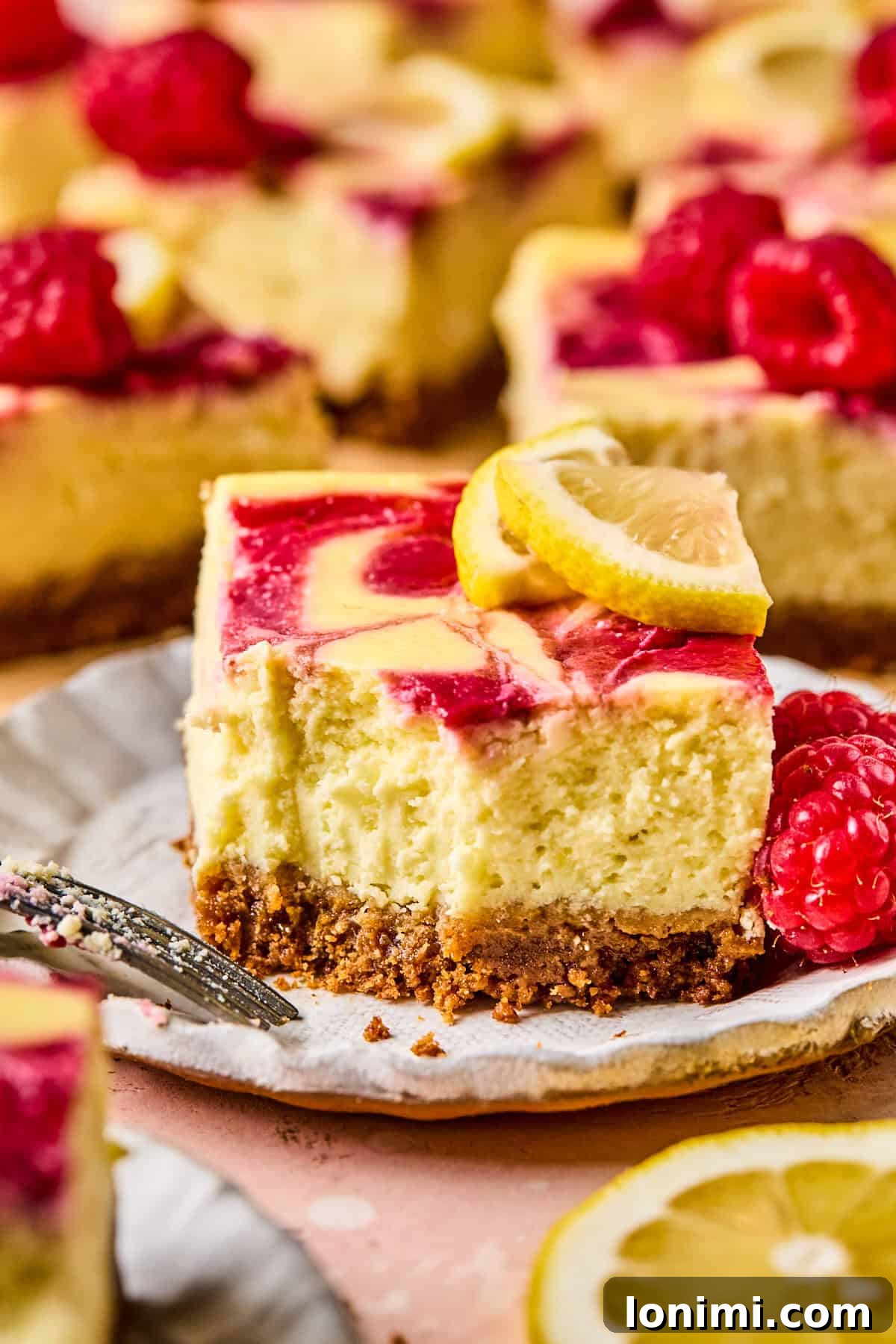 Close-up of a delectable slice of lemon raspberry cheesecake on a white plate. The cheesecake features a beautiful swirled raspberry topping and a perfectly golden graham cracker crust. It's elegantly garnished with fresh raspberries and a thin lemon slice, with a fork resting beside it, and other servings visible in the soft background.