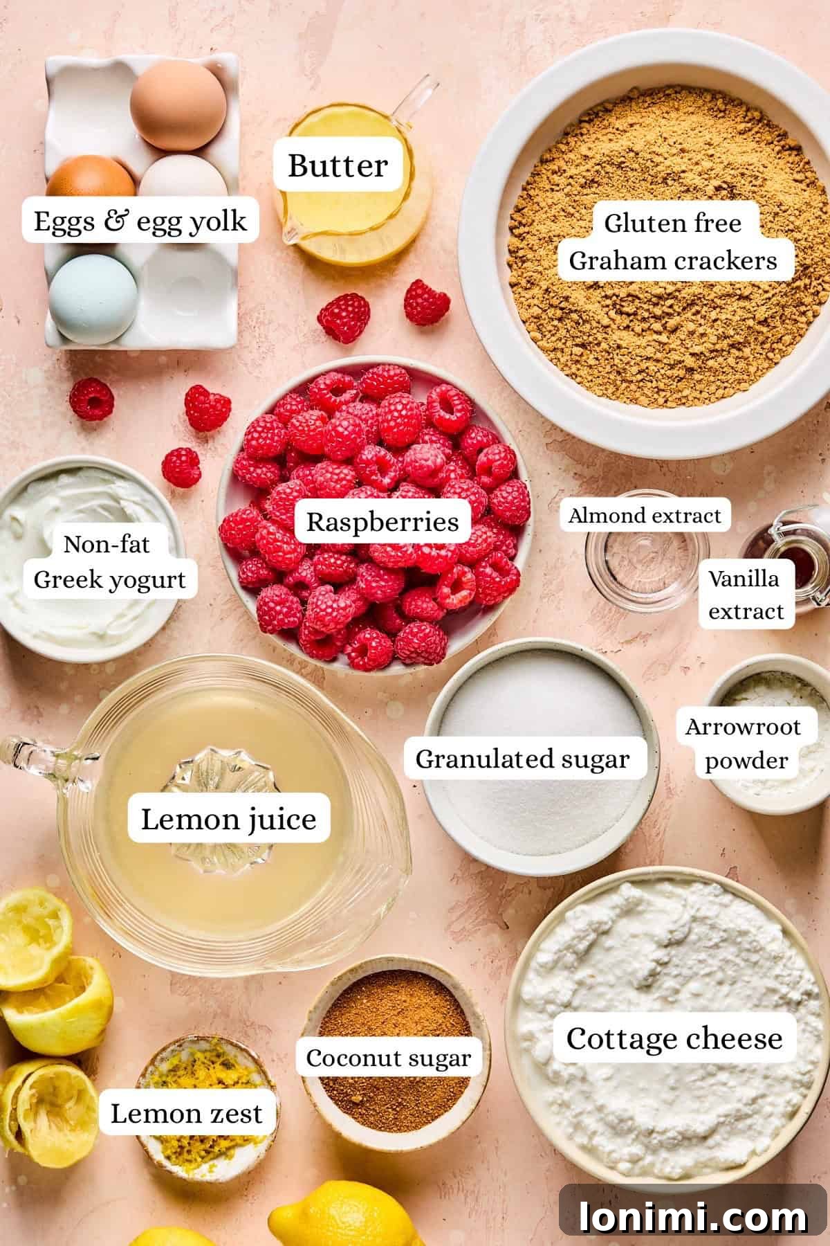 A beautifully arranged flat lay of all the essential ingredients needed for baking, including fresh eggs, melted butter, gluten-free graham crackers, vibrant raspberries, almond extract, vanilla extract, various sugars, fresh lemon juice and zest, Greek yogurt, arrowroot powder, and cottage cheese, all artfully displayed against a soft pink background.