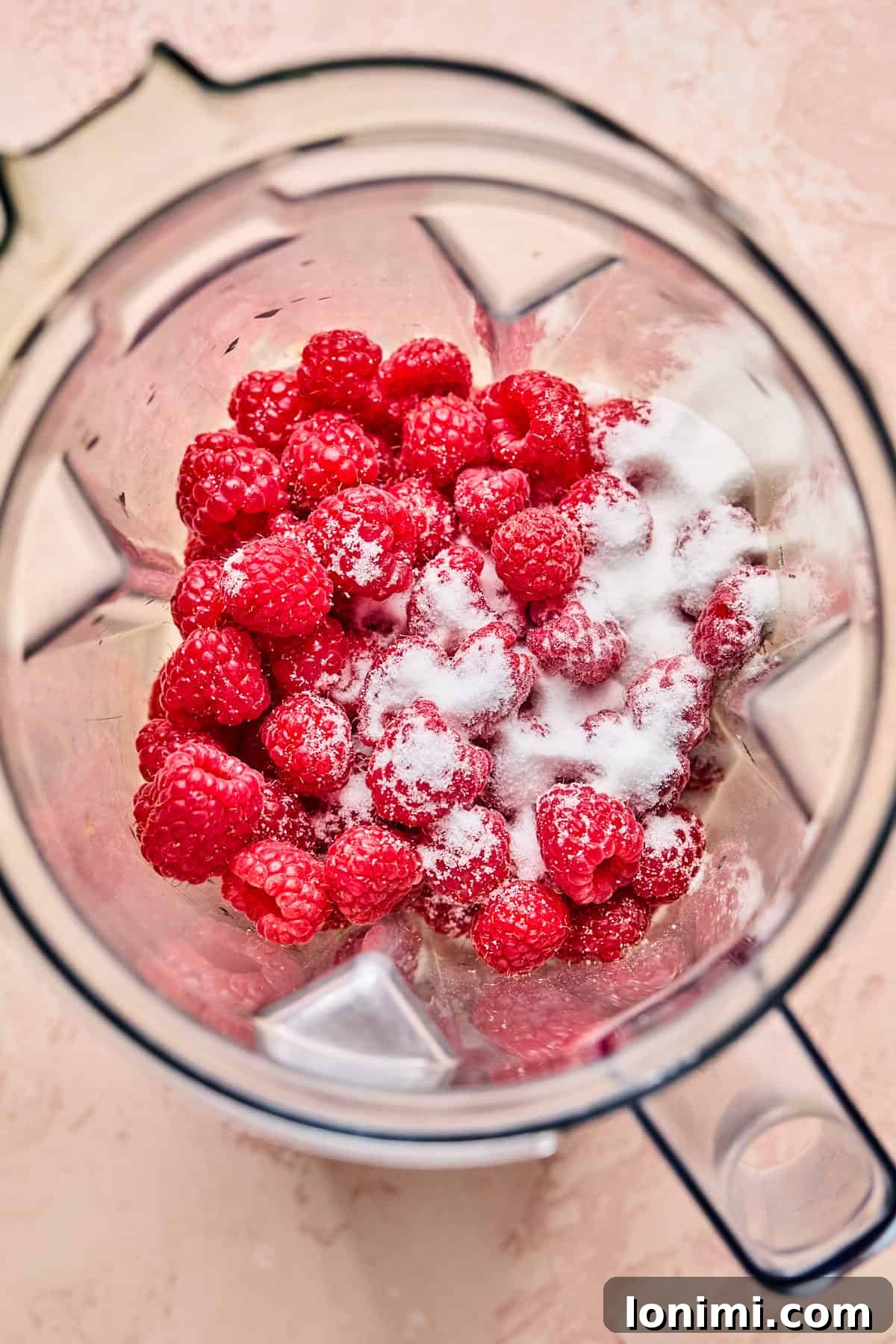 A top view of a blender containing fresh raspberries, some sprinkled with white sugar, against a light pink background, ready to be pureed for the cheesecake swirl.