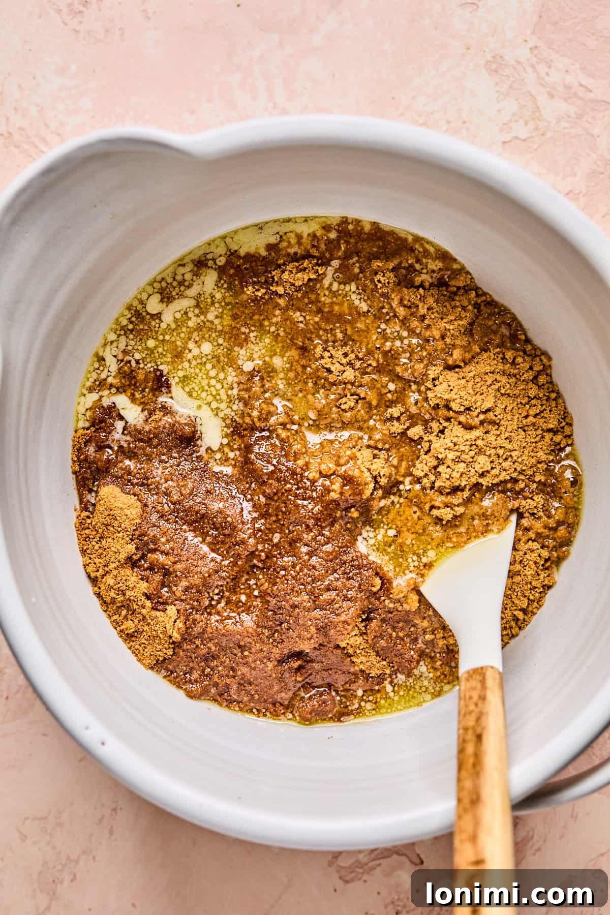 A mixing bowl containing a rich mixture of graham cracker crumbs, coconut sugar, and melted butter, with a wooden spoon resting on the edge, all on a pale pink textured surface.