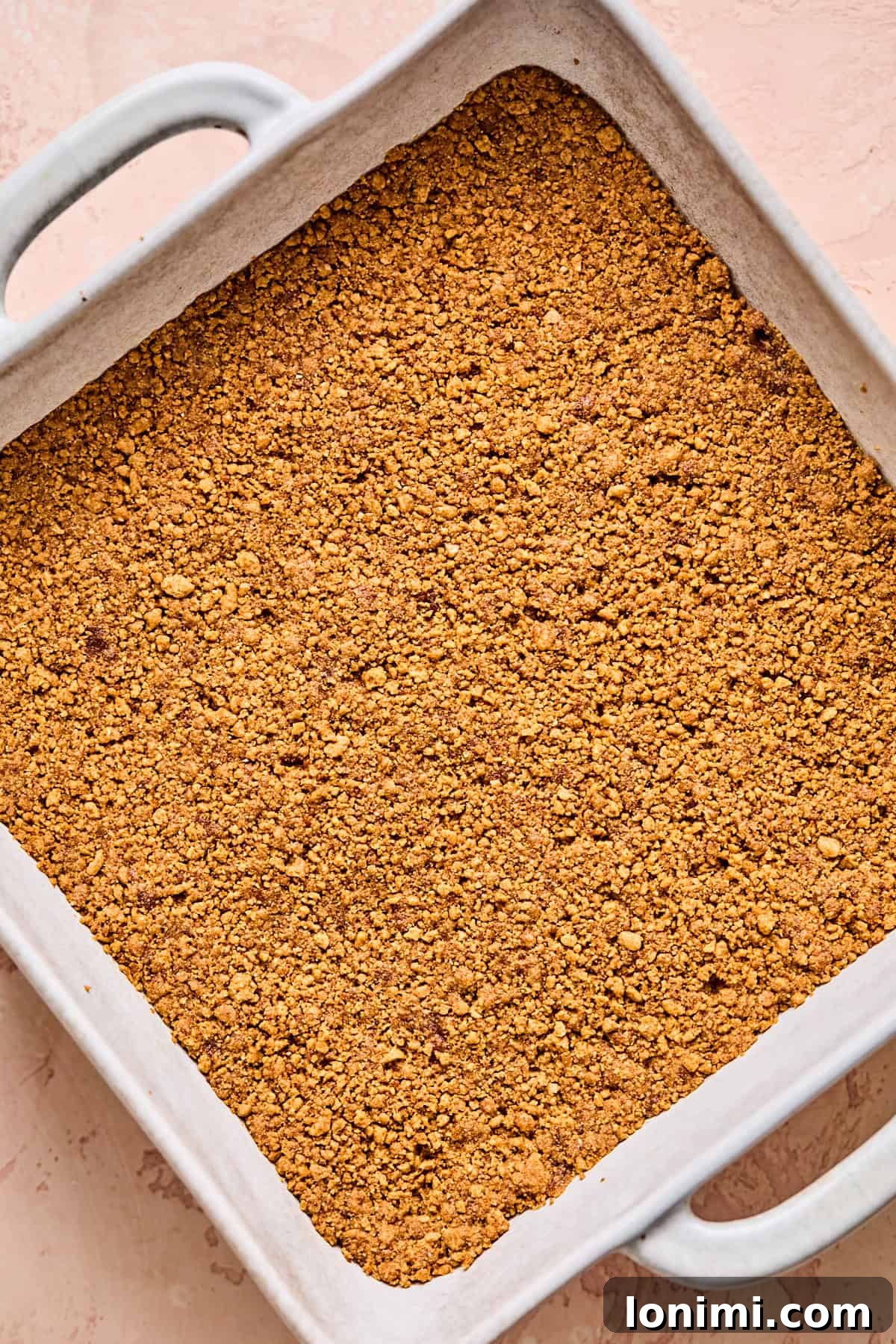 A square baking dish expertly lined with parchment paper and filled with a firm, even layer of golden graham cracker crust, ready for baking.