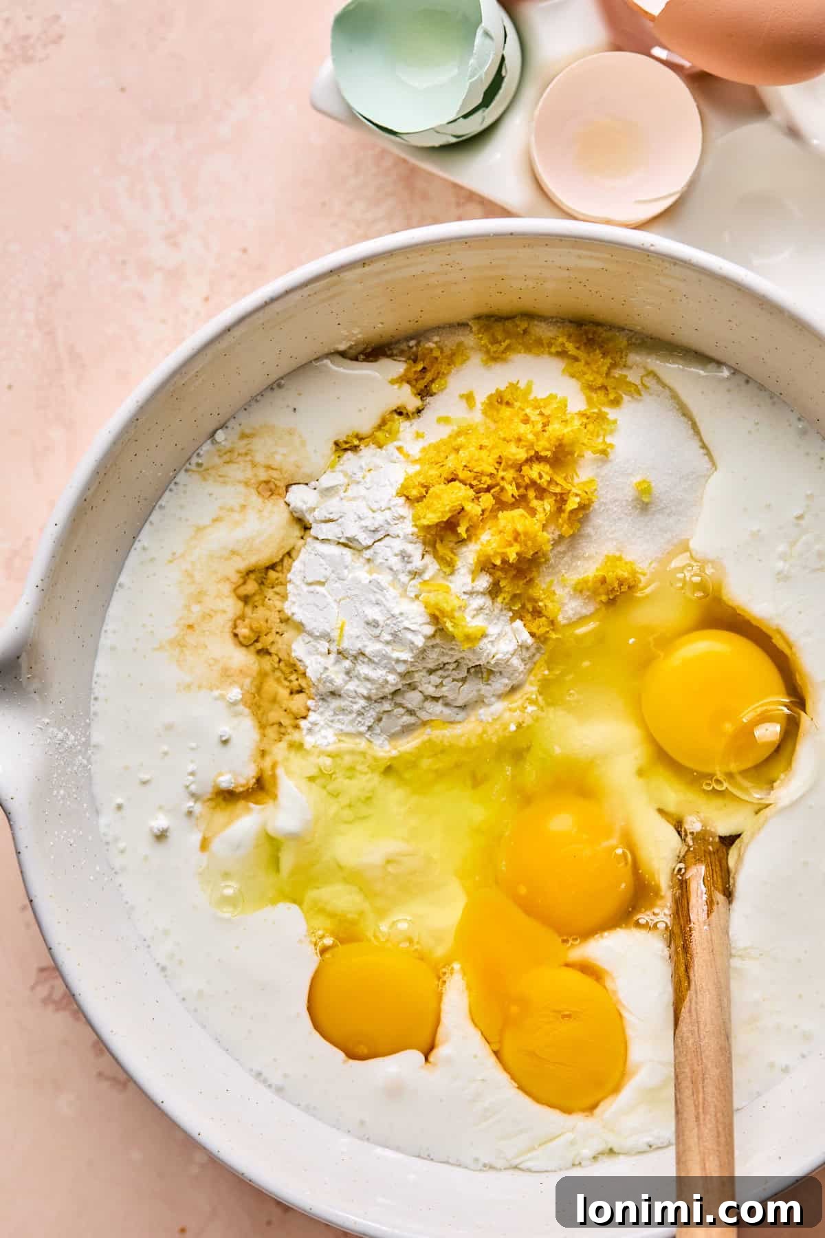 A large mixing bowl filled with all the ingredients for the protein cheesecake filling—creamy blended cottage cheese, Greek yogurt, sugars, arrowroot powder, eggs, lemon zest and juice, and extracts—ready to be combined.