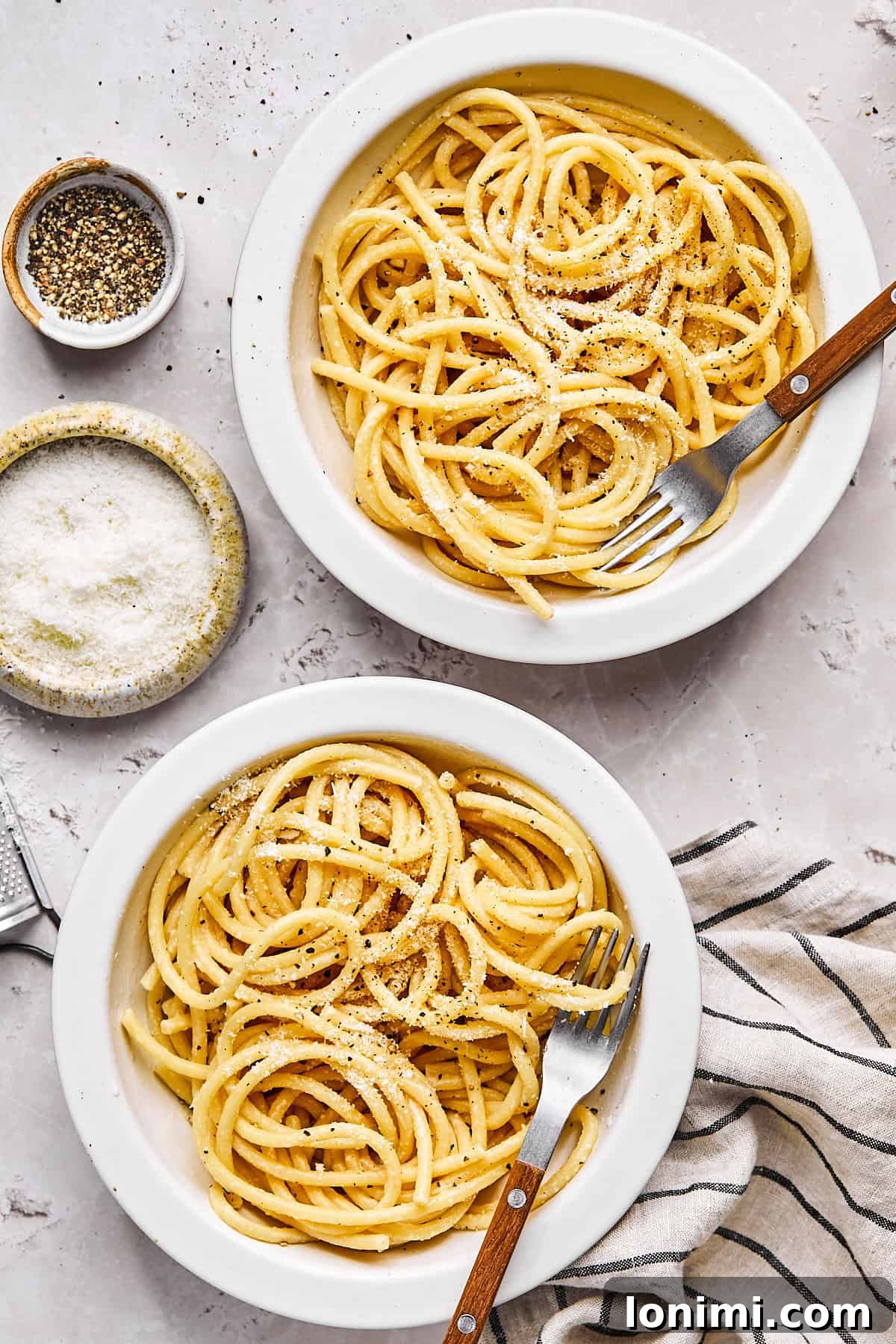 two bowls of gluten-free bucatini cacio e pepe next to a small dish of cheese and cracked pepper