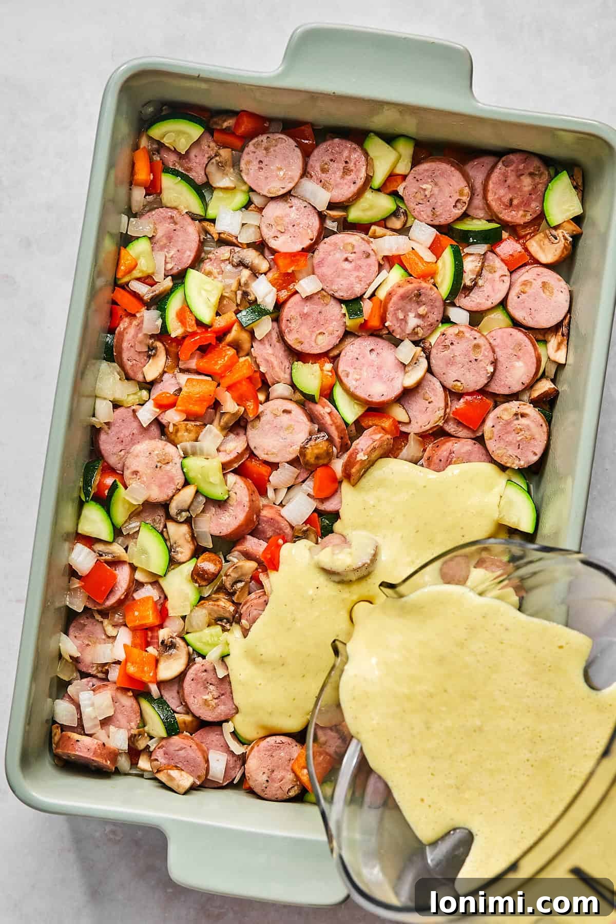 Casserole dish filled with sautéed veggies and sausage as blended egg mixture is poured on top